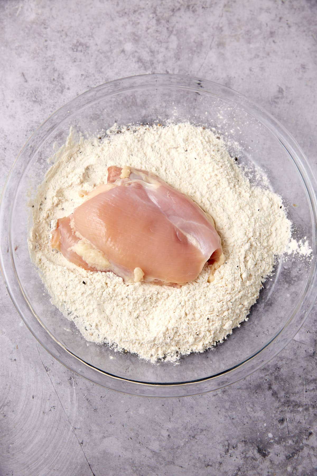 chicken thigh being dredged in flour in a glass bowl.