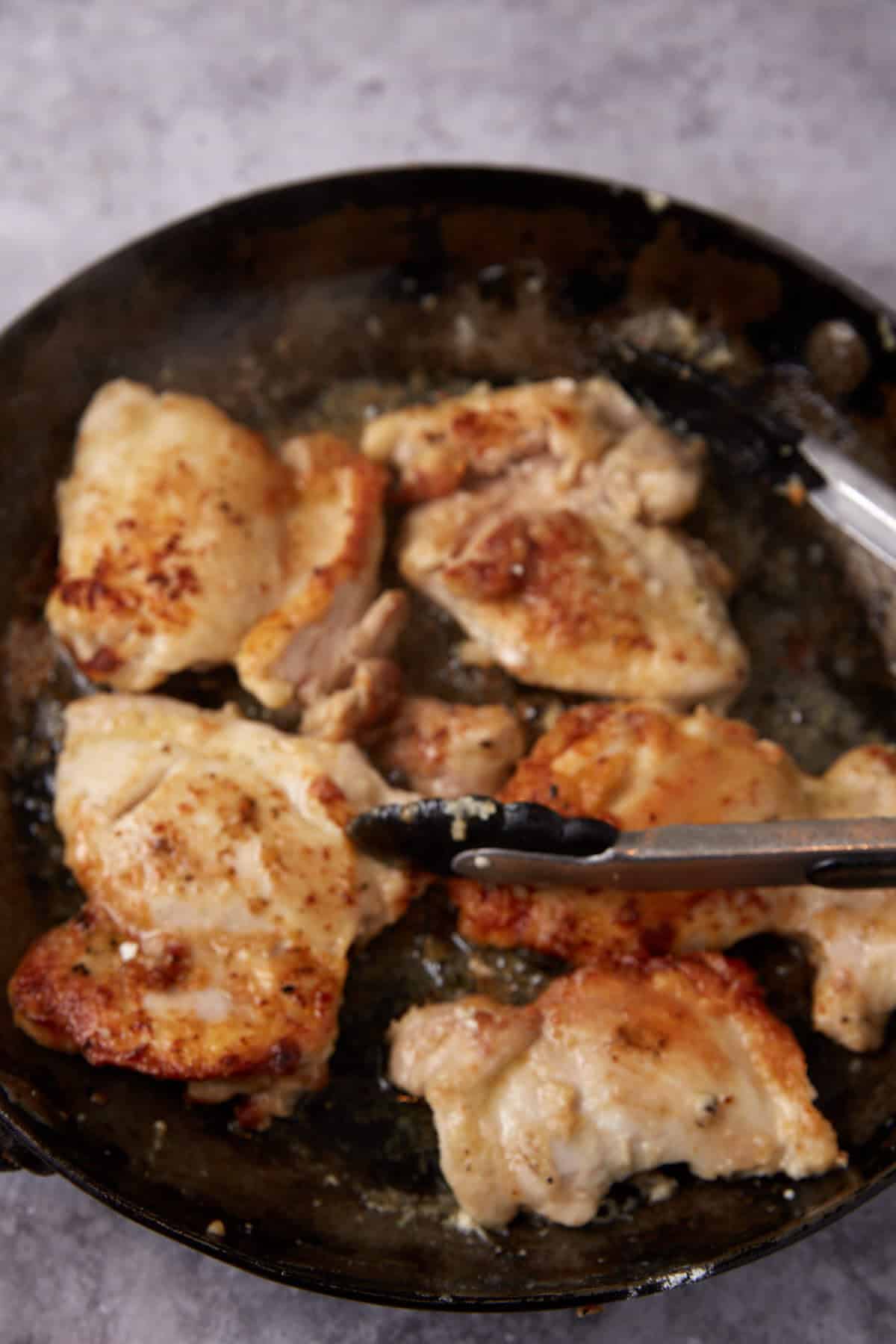chicken thighs being cooked in a skillet with tongs.