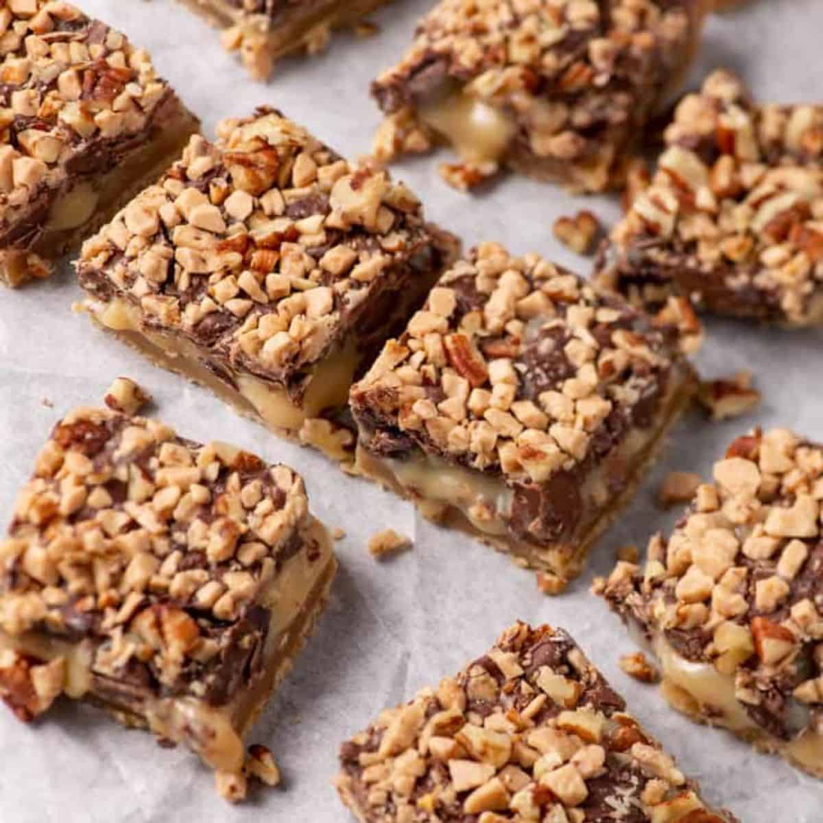A close-up image of several squares of Shortbread Toffee Bars, featuring a base of crumbly shortbread crust, a gooey caramel center, and a top layer covered in chopped nuts. The bars are arranged on a sheet of white parchment paper.