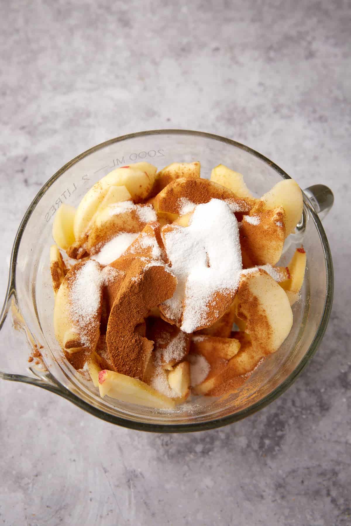 Sliced apples in a glass mixing bowl topped with cinnamon and sugar, ready to become a delicious apple crisp with oatmeal topping, placed on a gray countertop.