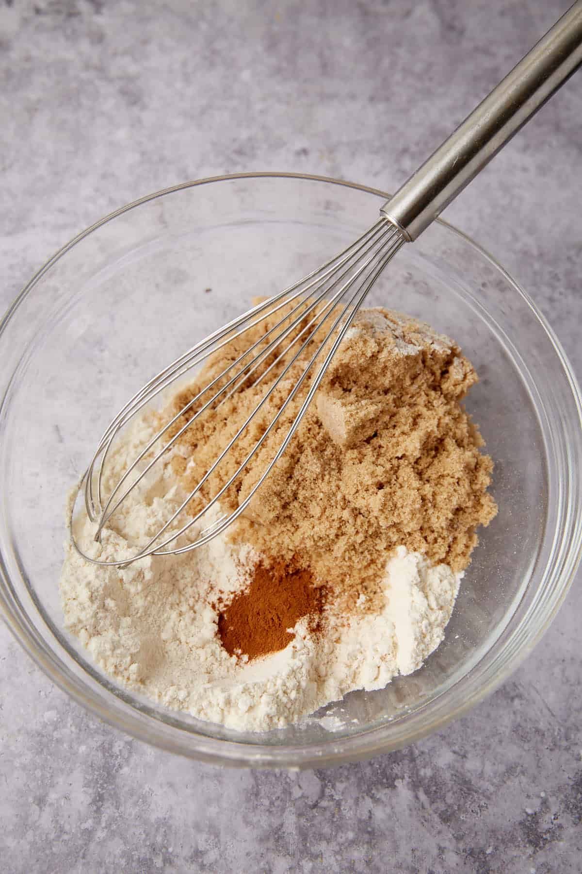 A glass bowl containing flour, brown sugar, and ground cinnamon, with a metal whisk resting on top—ingredients ready for mixing the perfect apple crisp with oatmeal on a gray countertop.