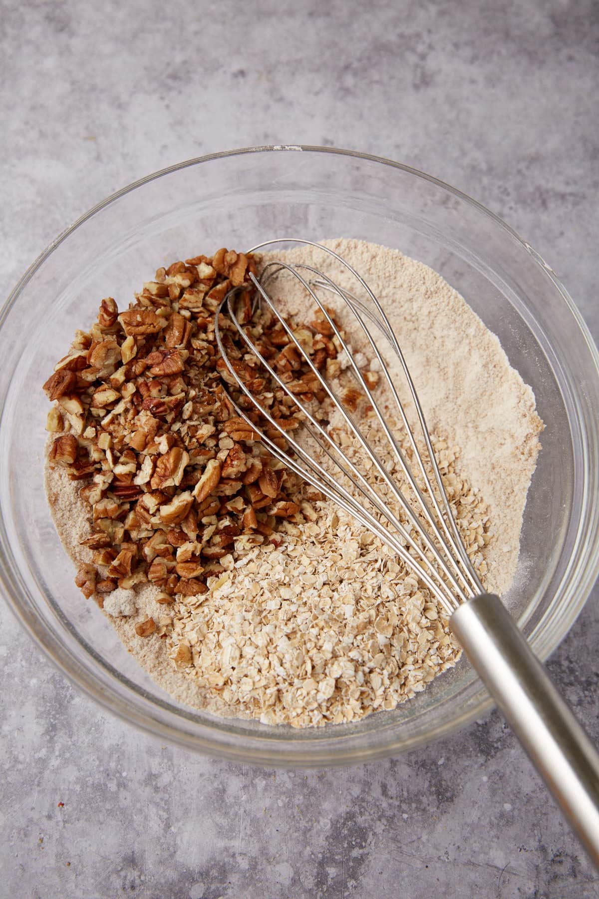 A glass mixing bowl containing flour, oats, and chopped pecans, ready for an Apple Crisp with Oatmeal Topping, with a metal whisk resting inside sits on a gray countertop.