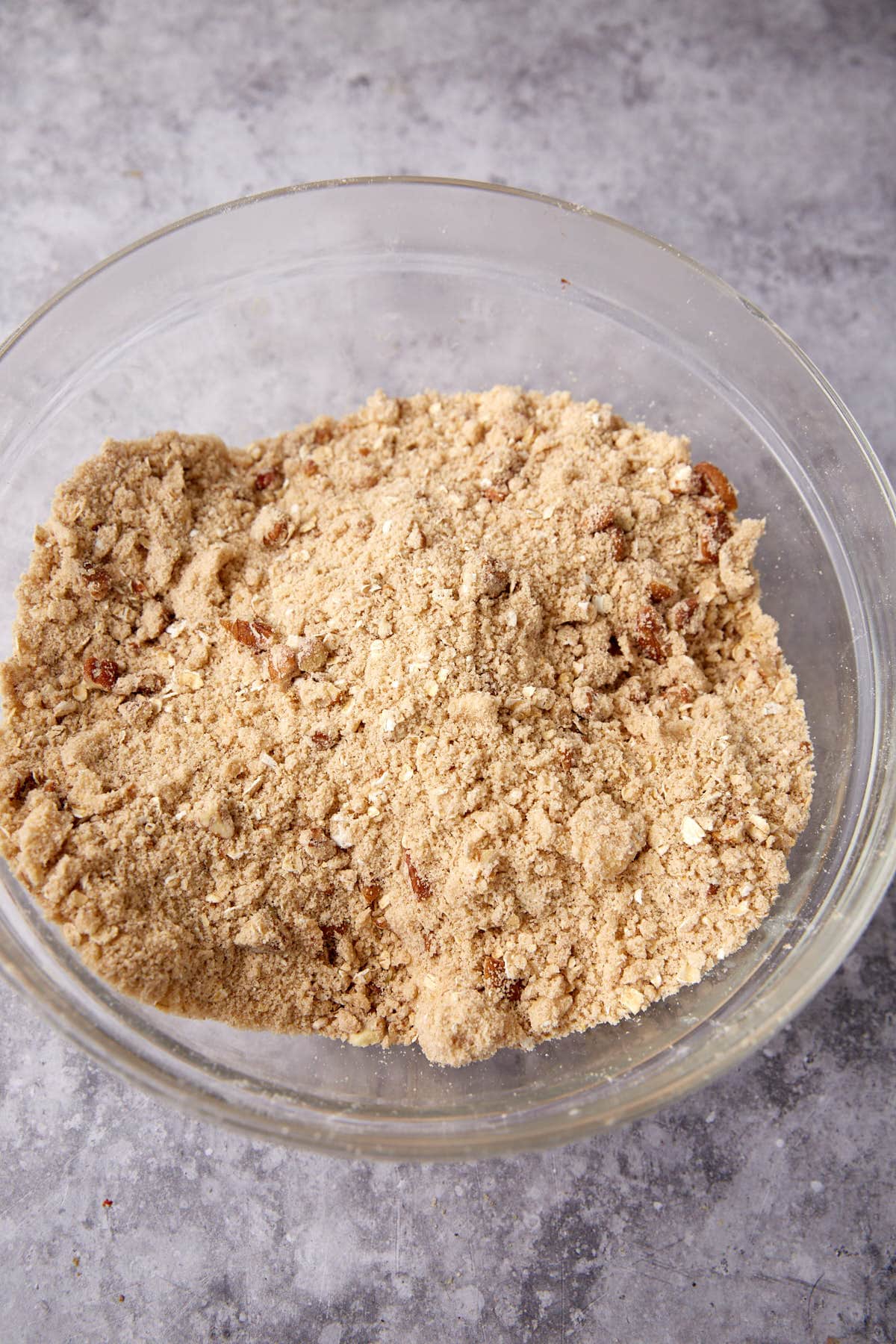 A glass bowl filled with a crumbly mixture of flour, sugar, and oats sits on a gray textured surface—perfect for making an oatmeal apple crisp or classic Apple Crisp with Oatmeal Topping.