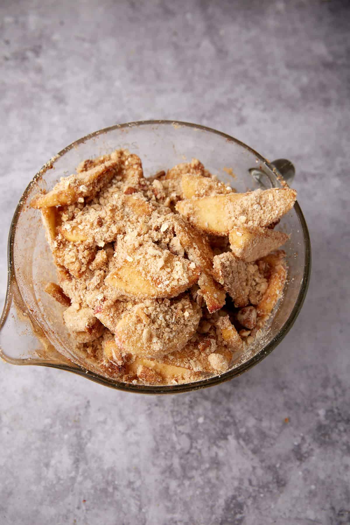 A glass mixing bowl filled with sliced apples coated in a crumbly brown sugar and cinnamon mixture, perfect for making a classic apple crisp with oatmeal topping, sits on a light gray countertop.