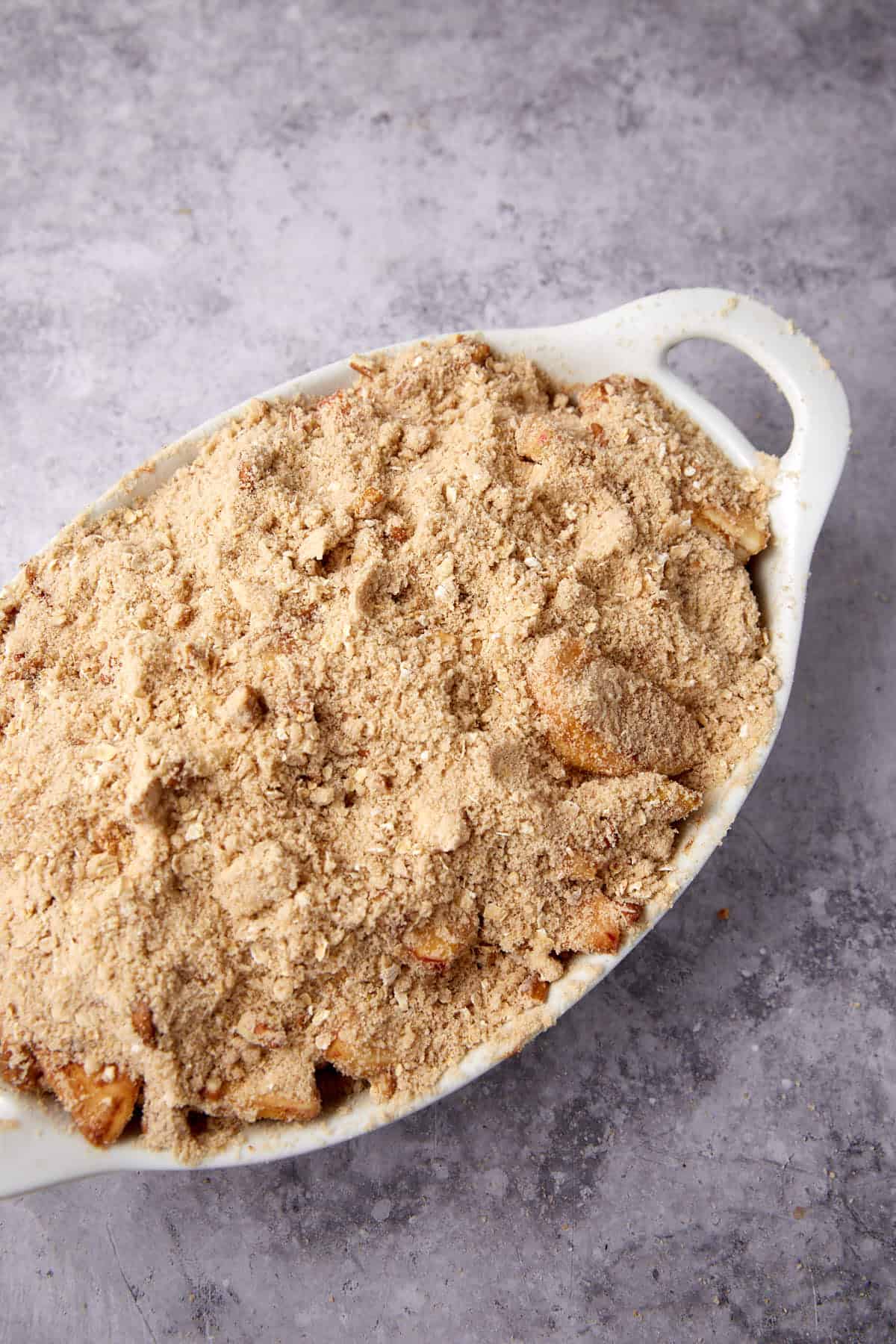 An oval white baking dish filled with oatmeal apple crisp, topped with a generous layer of crumbly, golden-brown streusel, sits on a gray textured surface.