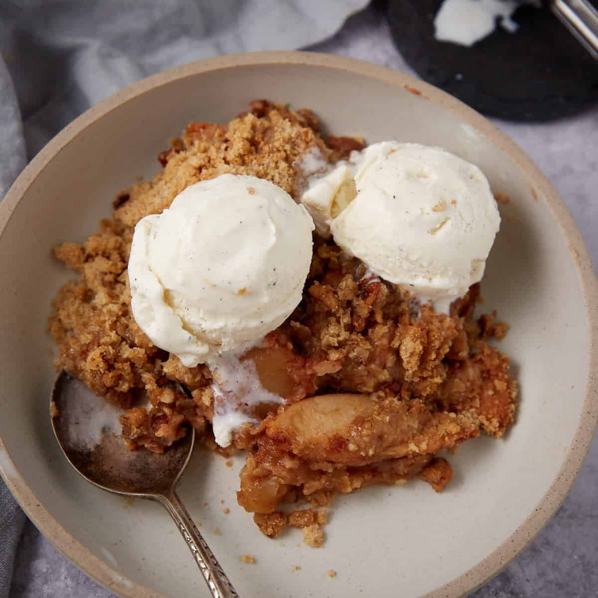 A bowl of apple crisp with oatmeal sits on a plate with a spoon, ready to eat. Two scoops of vanilla ice cream slowly melt over the warm crumble, making this oatmeal apple crisp an irresistible dessert.