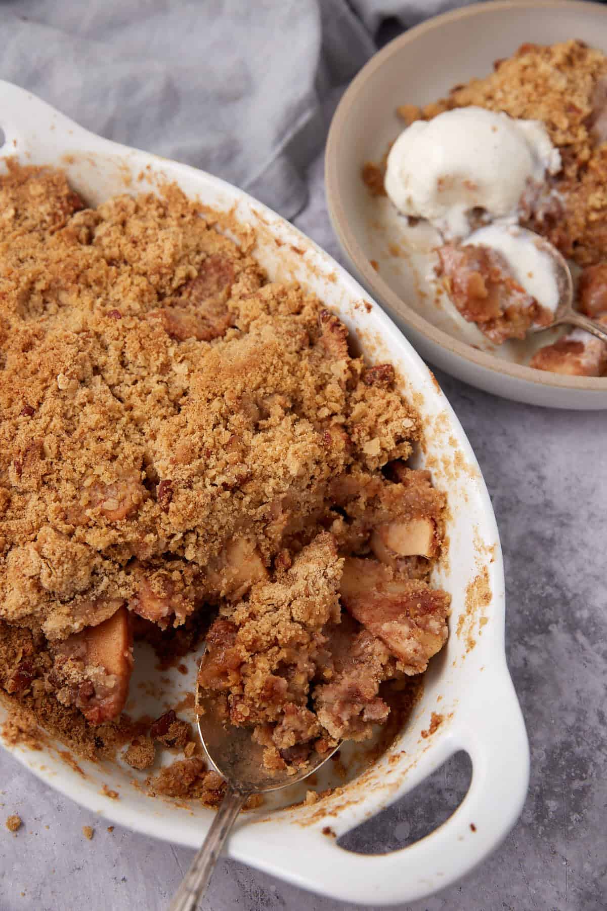 A white baking dish filled with apple crisp with oatmeal sits next to a bowl containing a serving of the dessert and two scoops of vanilla ice cream. A spoon rests in the baking dish, revealing spiced apples beneath the crumbly oatmeal topping.