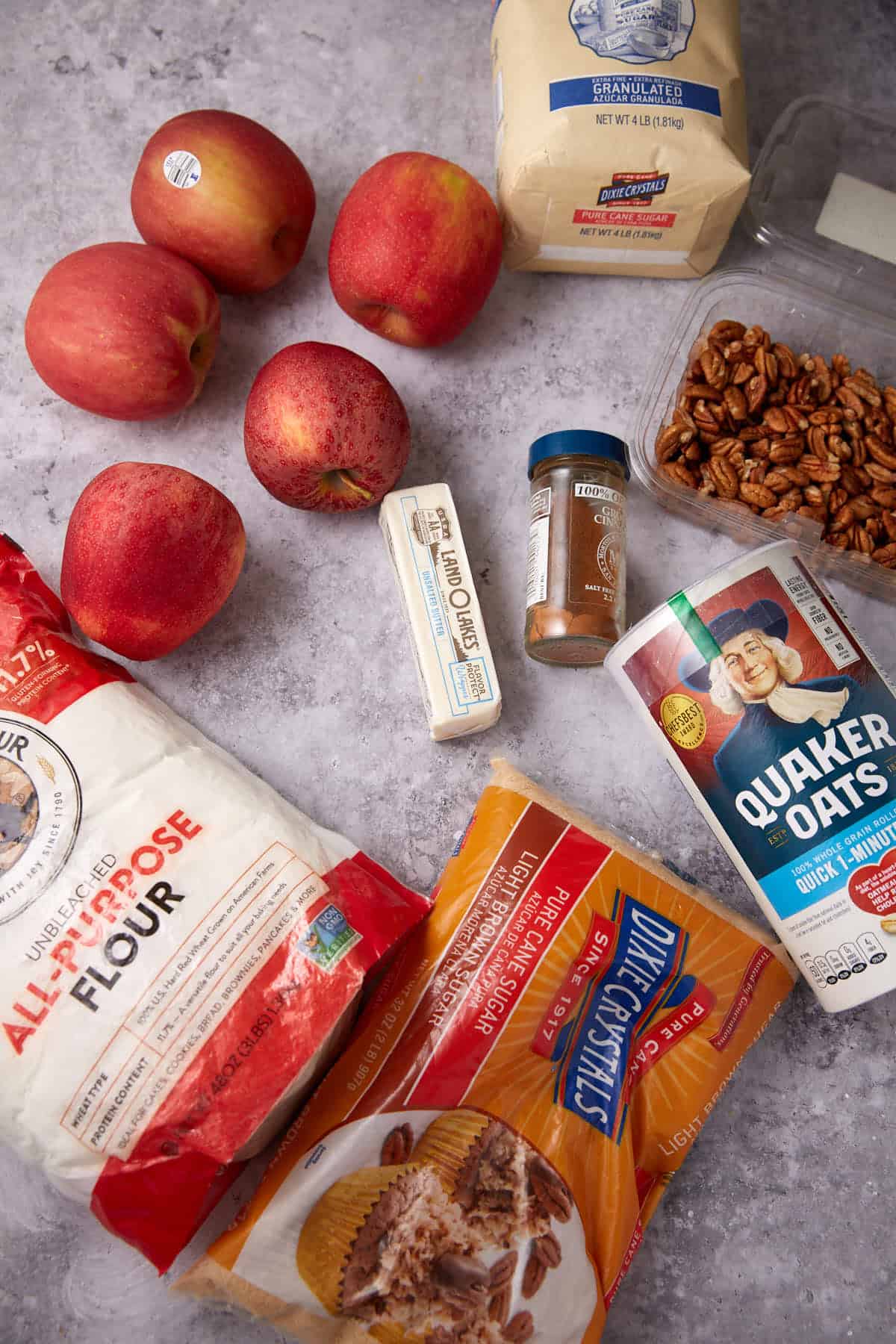 A top-down view of baking ingredients for an oatmeal apple crisp, featuring apples, flour, butter, pecans, cinnamon, sugar, and a container of Quaker oats arranged on a gray countertop.