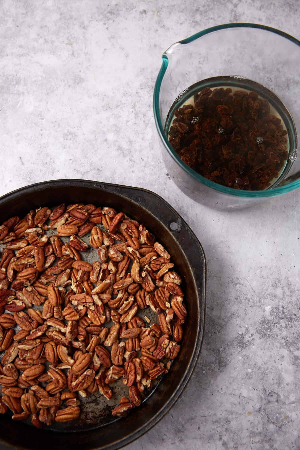 A round baking pan filled with pecan halves sits next to a glass measuring cup containing raisins soaking in water—perfect ingredients for crafting an Apple Cobbler with Sweet Potato Biscuits on a gray countertop.