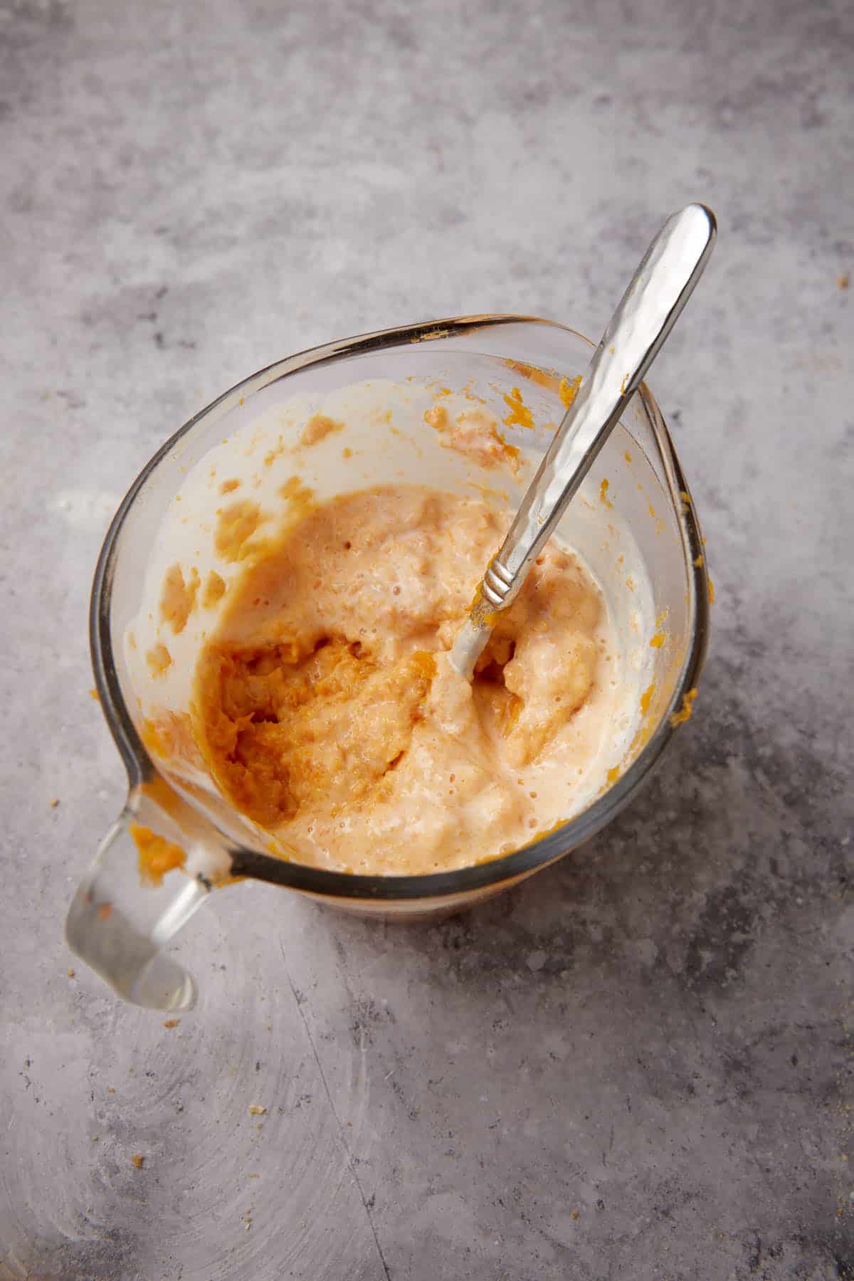 A glass measuring cup filled with a creamy orange mixture for Apple Cobbler with Sweet Potato Biscuits and a silver spoon, placed on a gray textured surface.