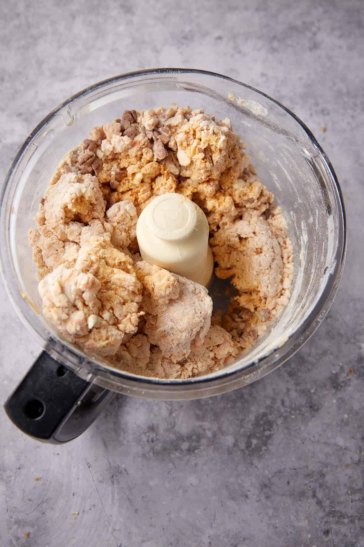 A food processor bowl containing partially mixed dough for Apple Cobbler with Sweet Potato Biscuits, with visible clumps and dry flour on the sides, set on a gray textured surface.