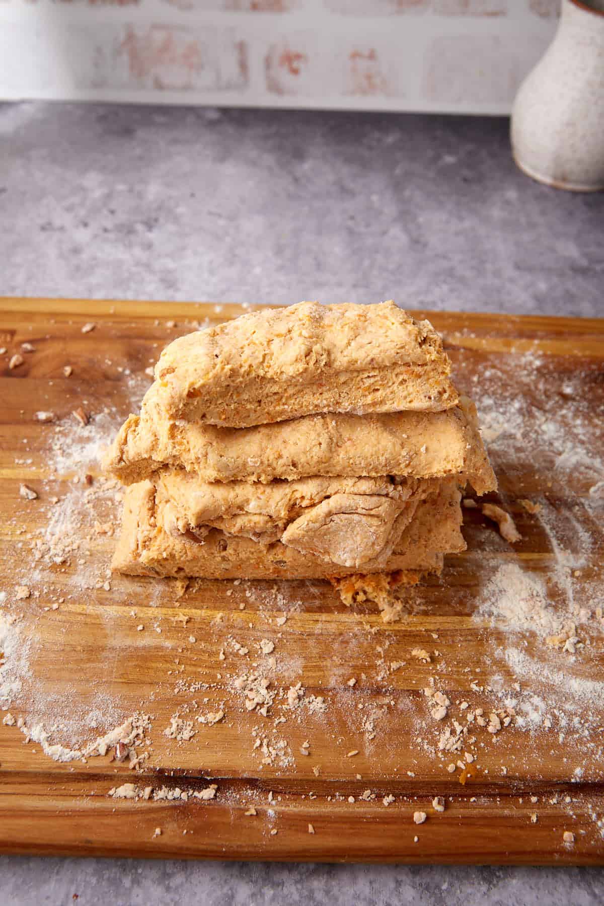 A stack of folded, rustic dough—perfect for Apple Cobbler with Sweet Potato Biscuits—rests on a floured wooden board, with flour scattered around. The background includes a stone countertop and part of a ceramic vessel.