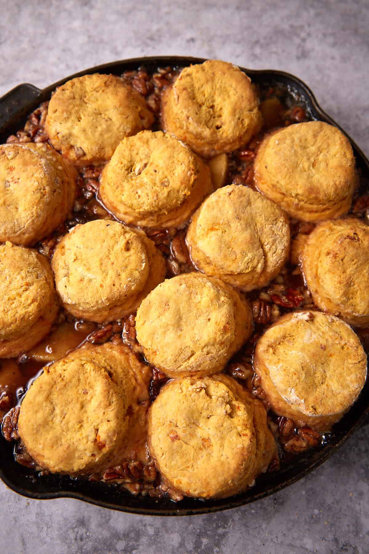A cast iron skillet filled with golden-brown sweet potato biscuits on top of a baked apple cobbler, featuring visible pecans and syrupy filling bubbling around the edges.