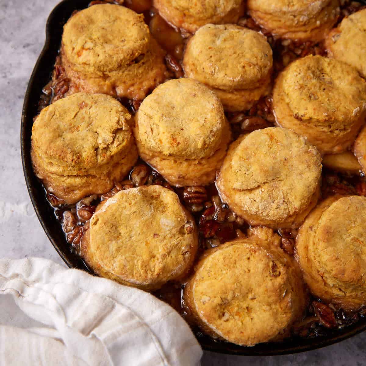 A cast iron skillet filled with golden brown apple cobbler with sweet potato biscuits sits atop a layer of chopped pecans and syrup, next to a white cloth napkin.