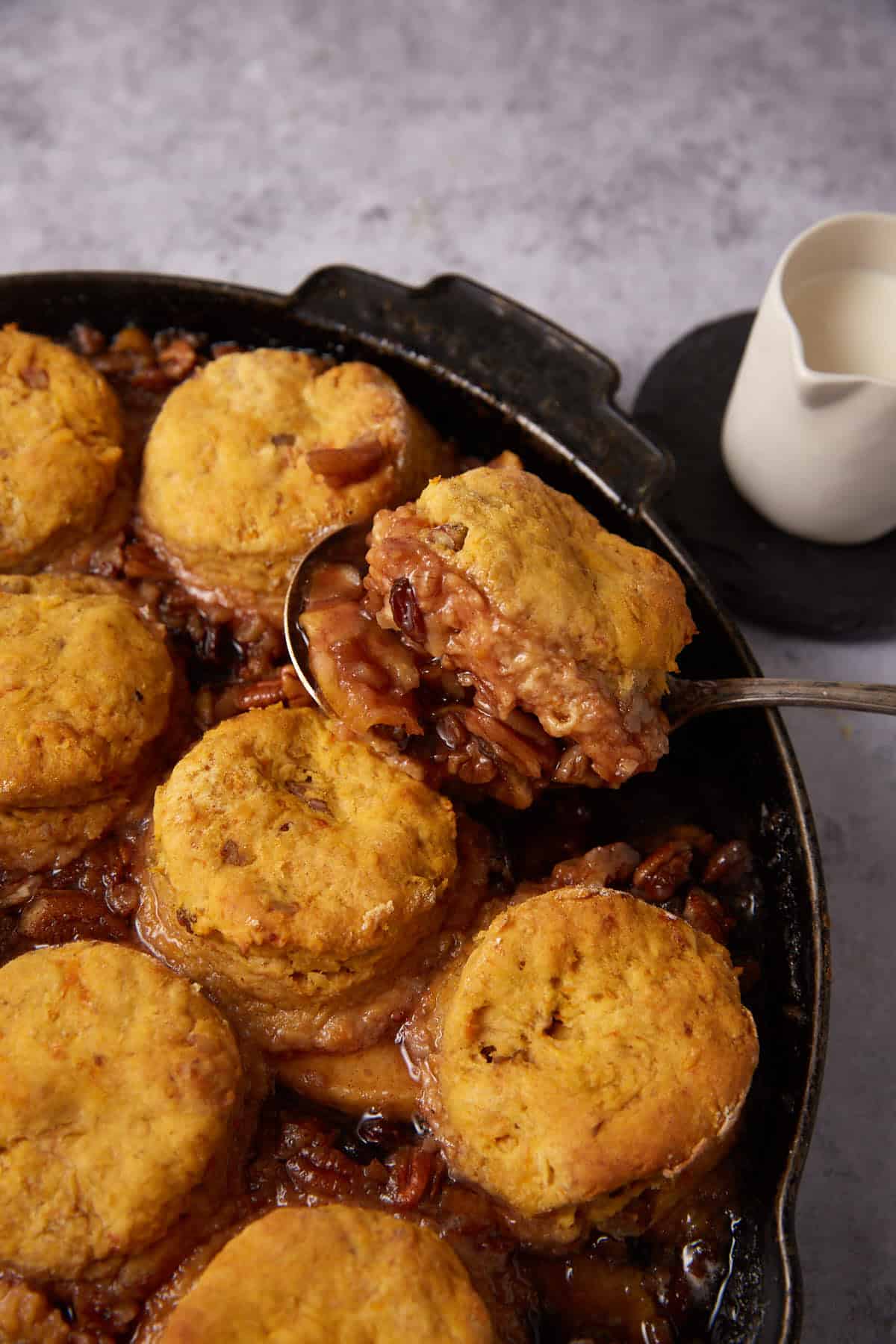A skillet filled with Apple Cobbler with Sweet Potato Biscuits atop a caramelized filling, being served with a spoon. A small white pitcher sits in the background on a dark coaster.