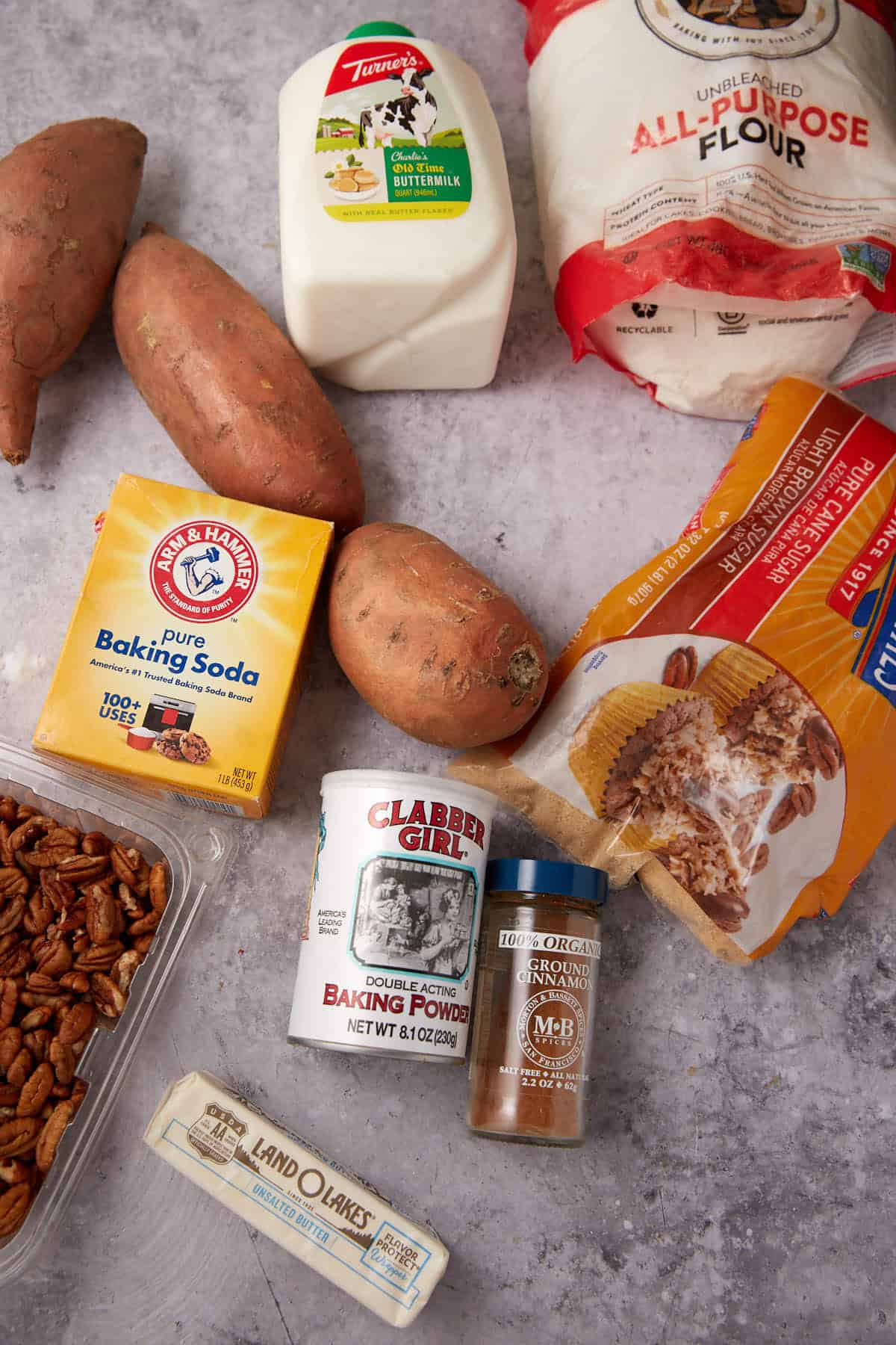 A flat lay of baking ingredients, perfect for making Apple Cobbler with Sweet Potato Biscuits: sweet potatoes, all-purpose flour, milk, baking soda, baking powder, pecans, cinnamon, brown sugar, butter, and granulated sugar on a gray surface.