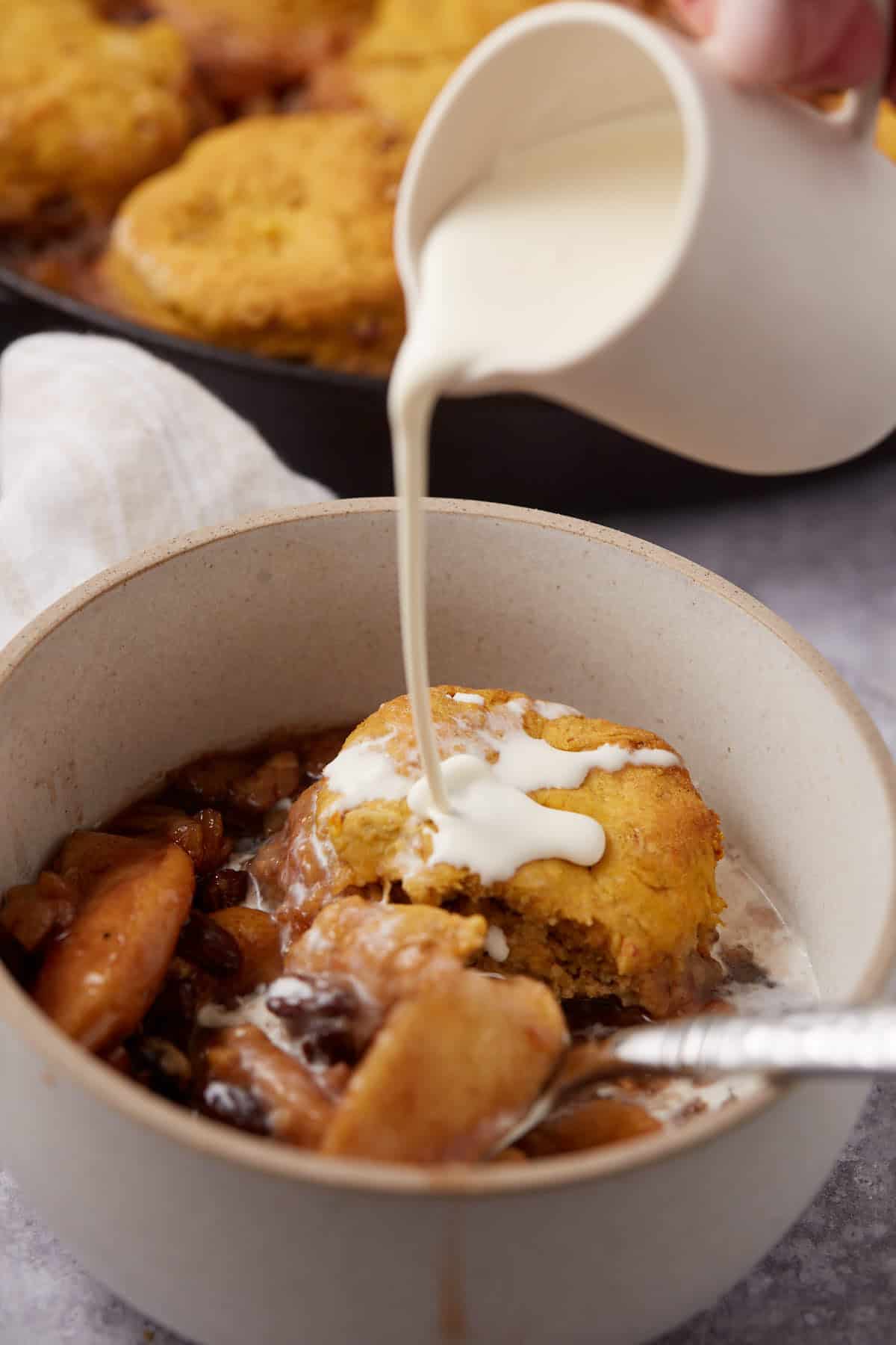 A bowl of Apple Cobbler with Sweet Potato Biscuits is topped with a biscuit as cream is poured from a small pitcher. A spoon rests in the bowl, and a baking dish appears in the background.