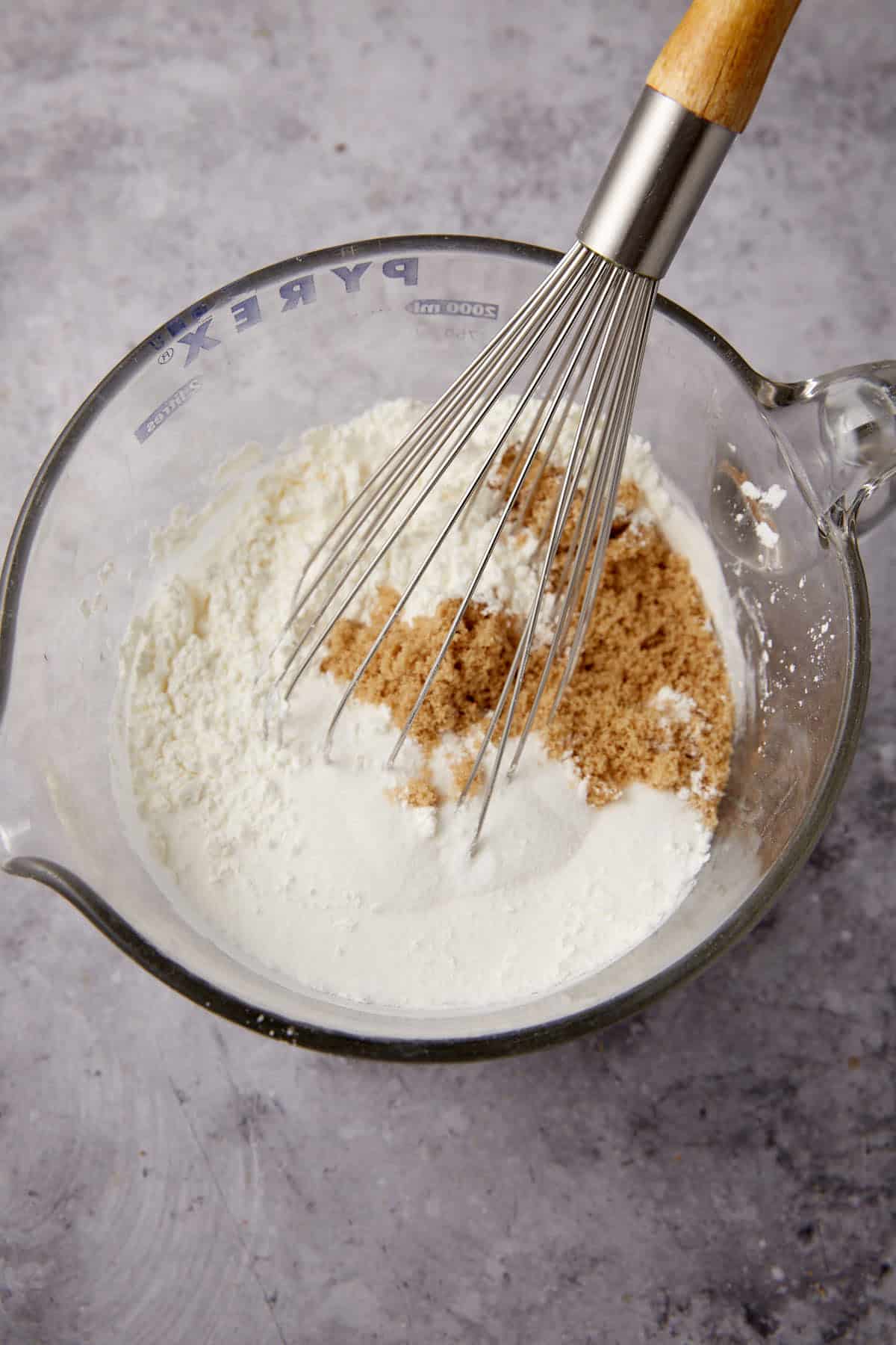 A glass measuring cup with flour, sugar, and brown sugar being mixed with a metal whisk on a gray countertop—perfect for preparing Apple Cobbler with Sweet Potato Biscuits.