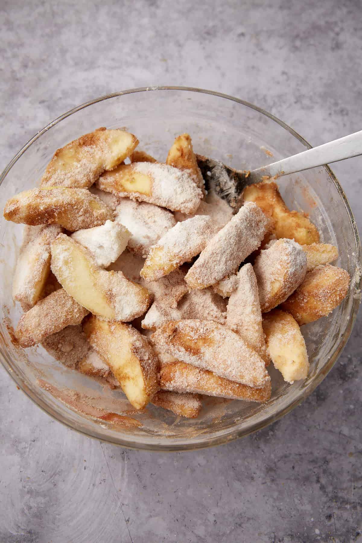 A glass bowl filled with sliced apples coated in a mixture of sugar and cinnamon, like the filling for Apple Cobbler with Sweet Potato Biscuits, with a metal spoon resting inside, on a gray textured surface.