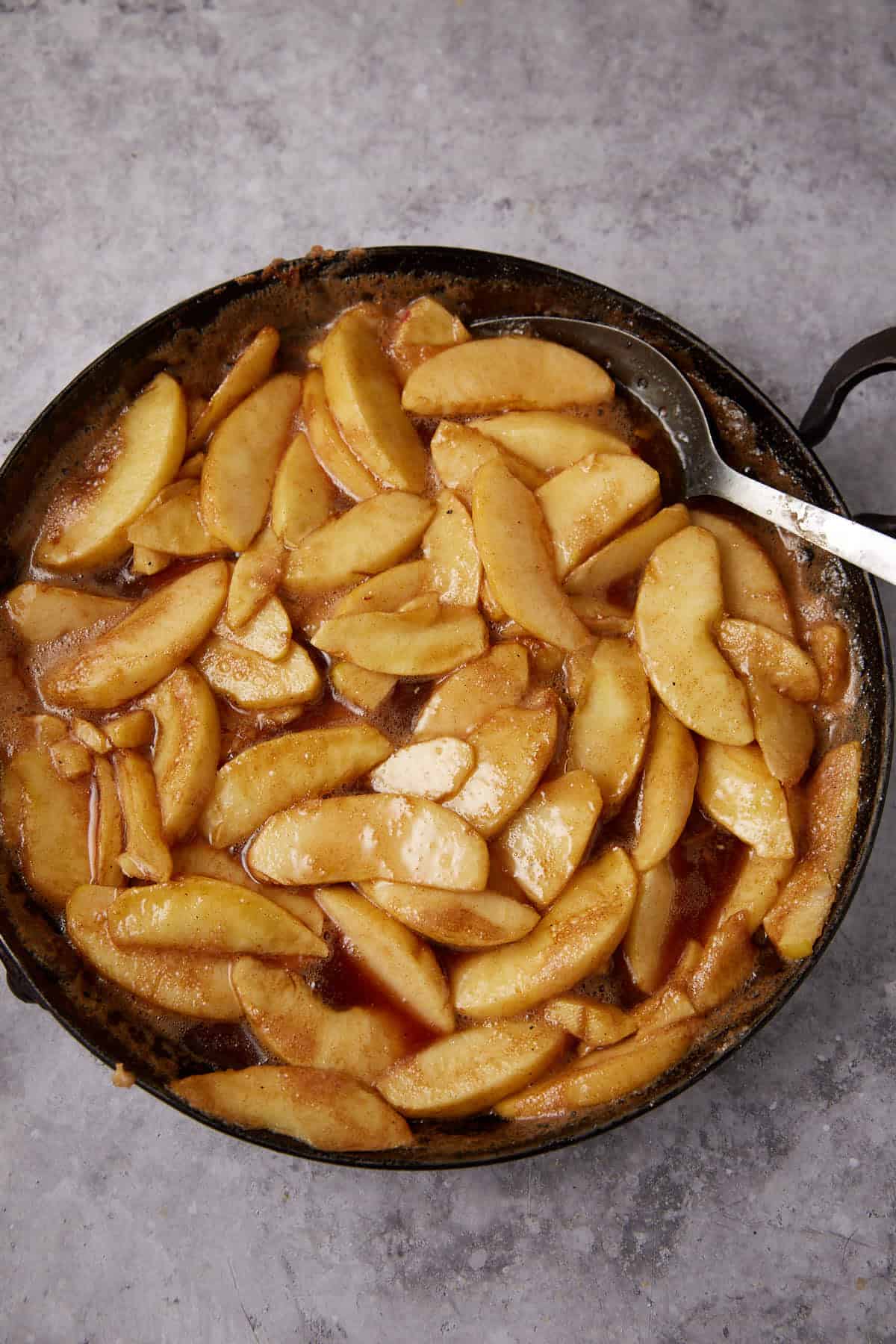 A pan filled with cooked, sliced apples coated in a glossy brown cinnamon sauce, inspired by Apple Cobbler with Sweet Potato Biscuits, with a metal serving spoon resting on the side. The background is a gray textured surface.