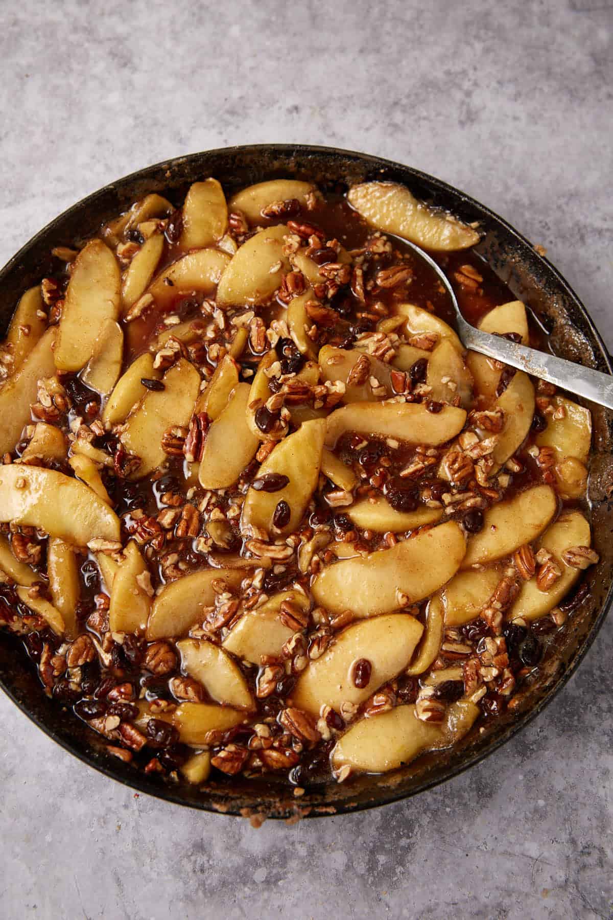 A skillet filled with sliced apples, chopped pecans, and raisins in a glossy brown sauce, perfect for serving alongside Apple Cobbler with Sweet Potato Biscuits, with a spoon resting inside on a gray textured surface.