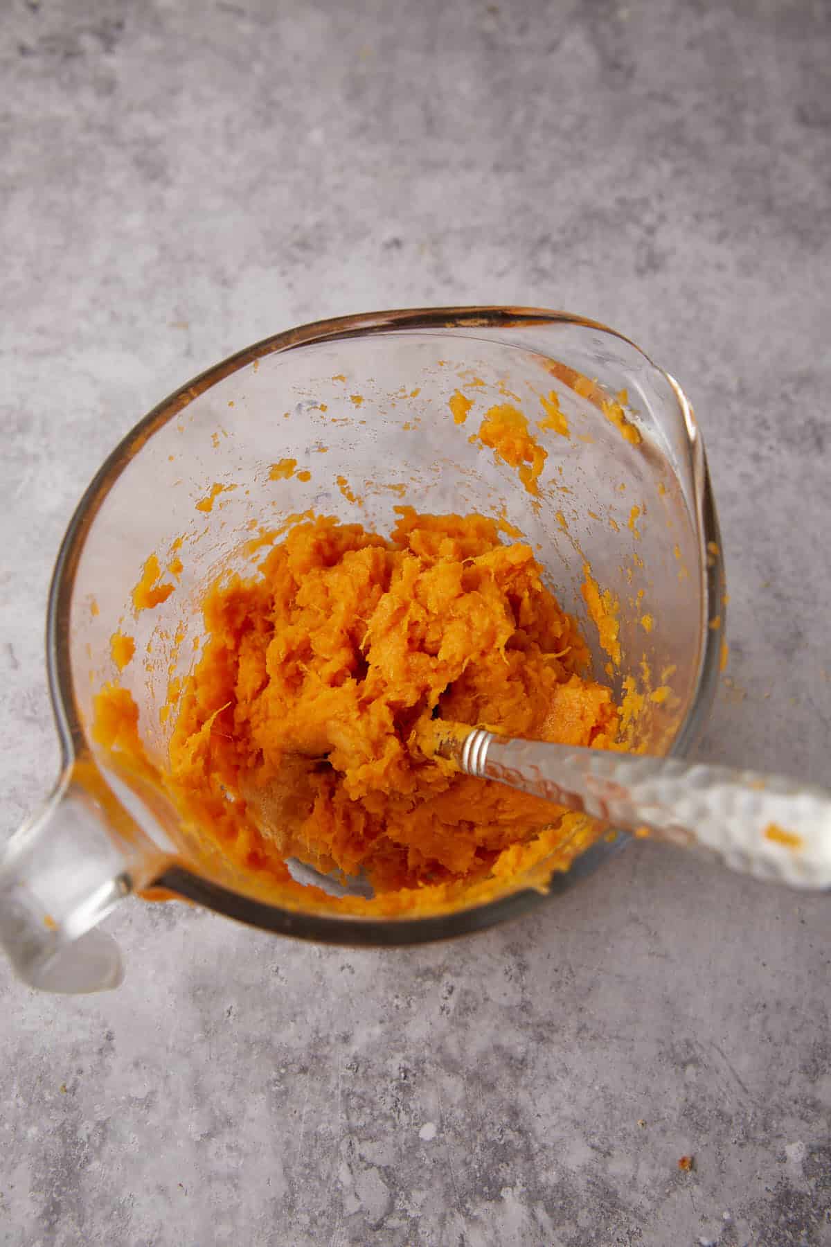 A glass mixing bowl containing mashed sweet potatoes with an ornate metal spoon, placed on a gray textured countertop, ready to be transformed into Apple Cobbler with Sweet Potato Biscuits.