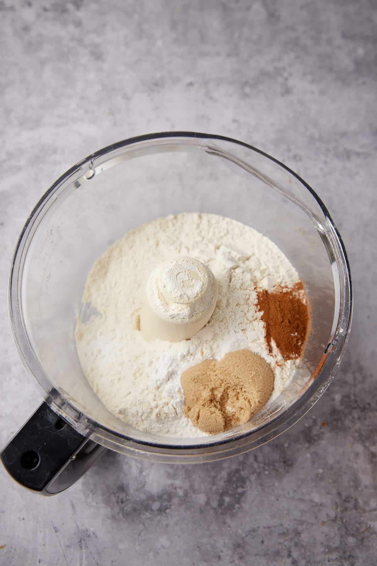 A food processor bowl filled with flour, brown sugar, and cinnamon—ingredients for Apple Cobbler with Sweet Potato Biscuits—sits on a gray countertop, ready to be mixed.