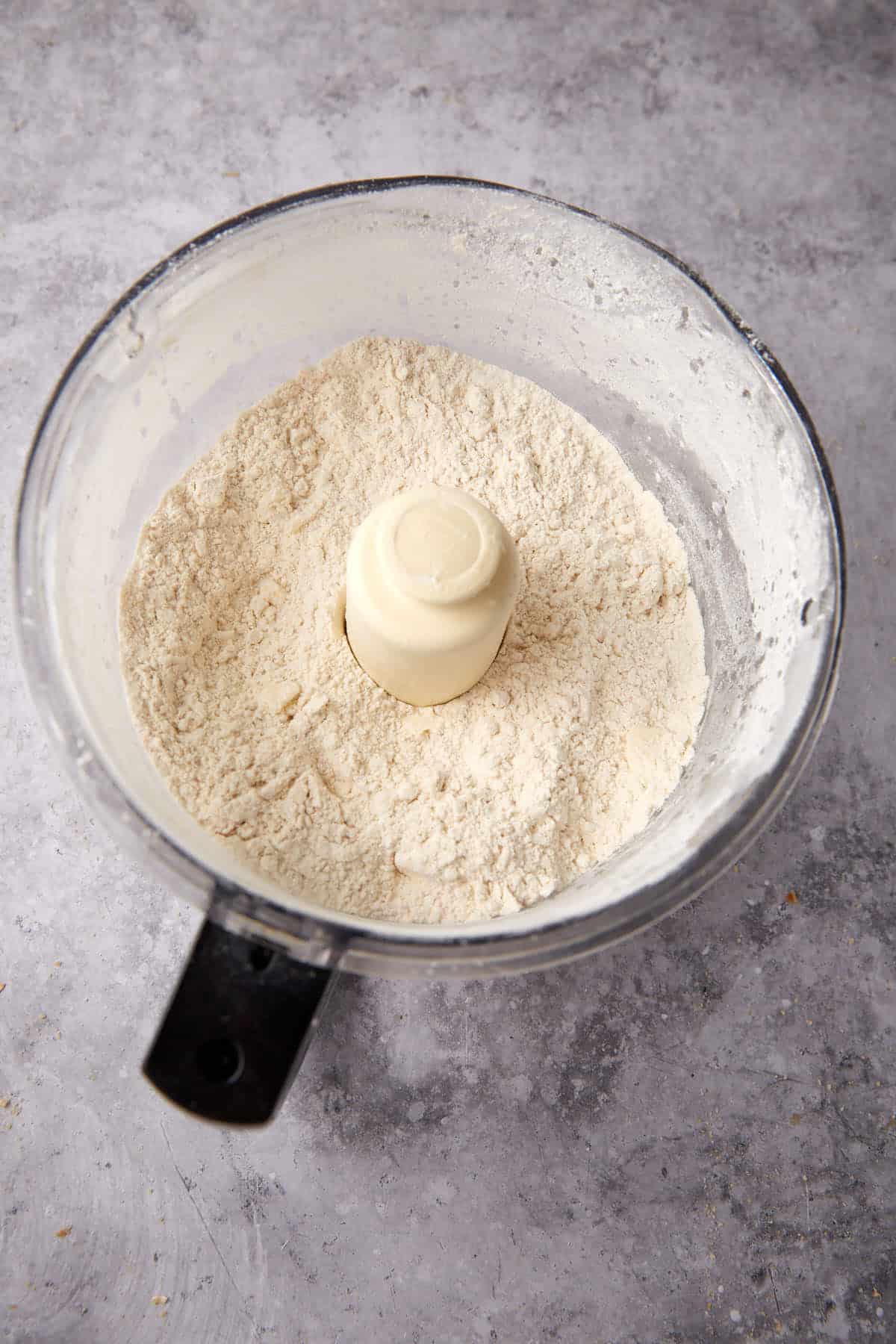 A food processor bowl filled with flour mixture for Apple Cobbler with Sweet Potato Biscuits sits on a gray countertop, viewed from above. The flour is finely mixed and surrounds the central blade attachment.
