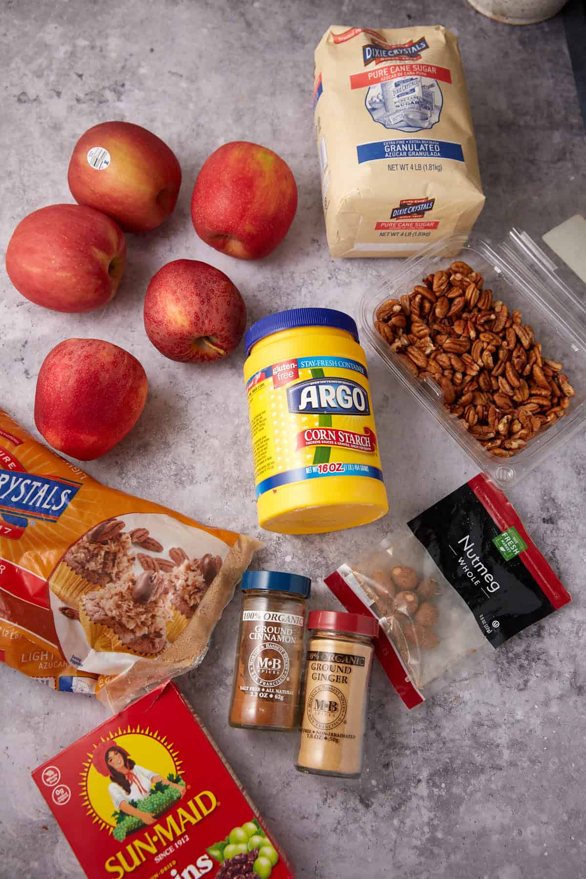 A flat lay of ingredients for Apple Cobbler with Sweet Potato Biscuits on a gray surface, featuring apples, flour, pecans, cornstarch, crystalized sugar, raisins, ground cinnamon and ginger, nutmeg, and whole nutmeg.