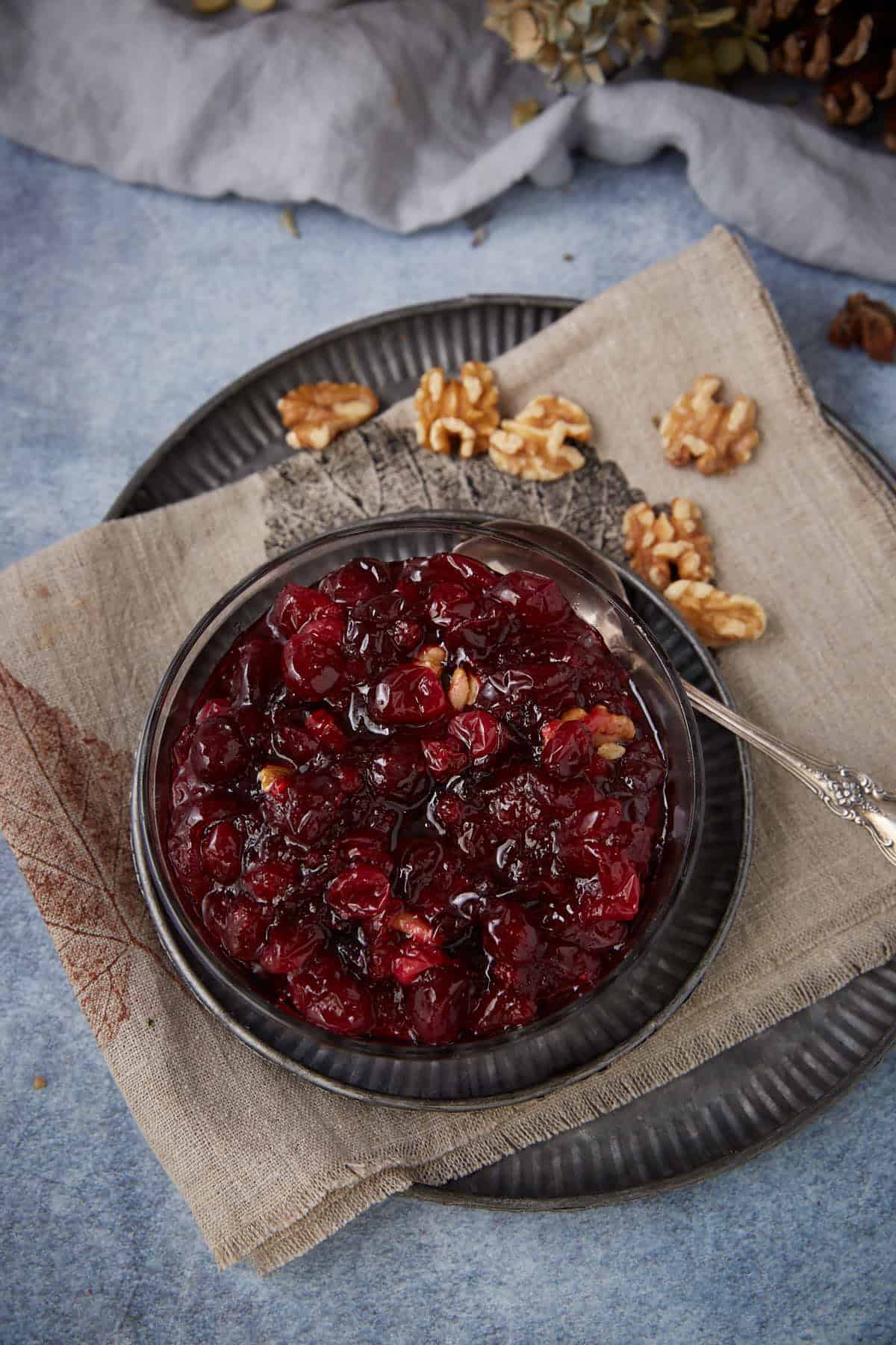 A bowl of homemade cranberry sauce with walnuts sits on a beige napkin on a dark plate, with a silver spoon and extra walnut pieces beside it on a blue and gray background.