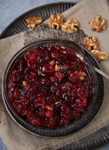 A glass bowl filled with chunky homemade cranberry sauce containing walnuts sits on a beige napkin atop a dark plate, with shelled walnut pieces scattered beside it. A silver spoon rests in the bowl.