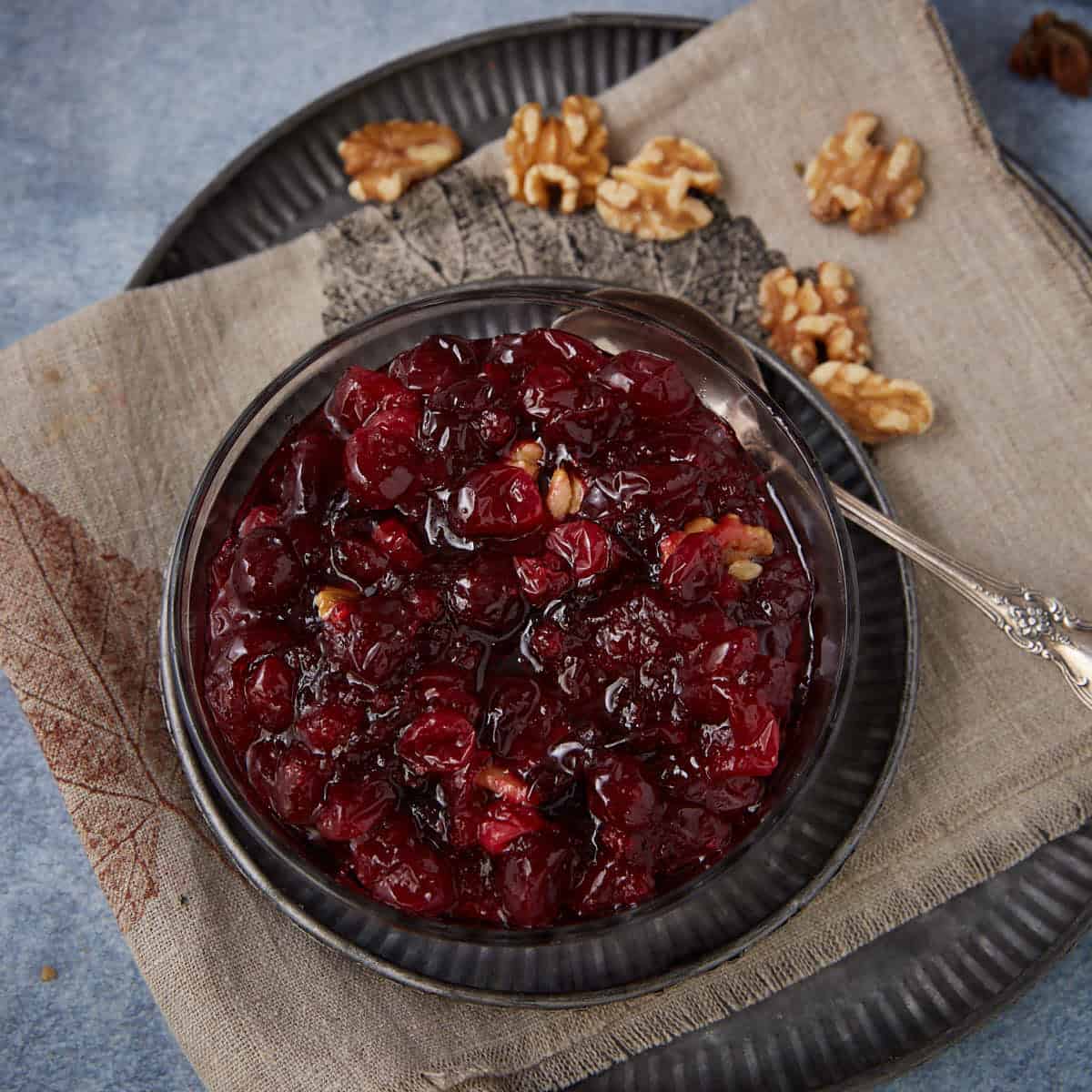 A glass bowl filled with chunky homemade cranberry sauce containing walnuts sits on a beige napkin atop a dark plate, with shelled walnut pieces scattered beside it. A silver spoon rests in the bowl.