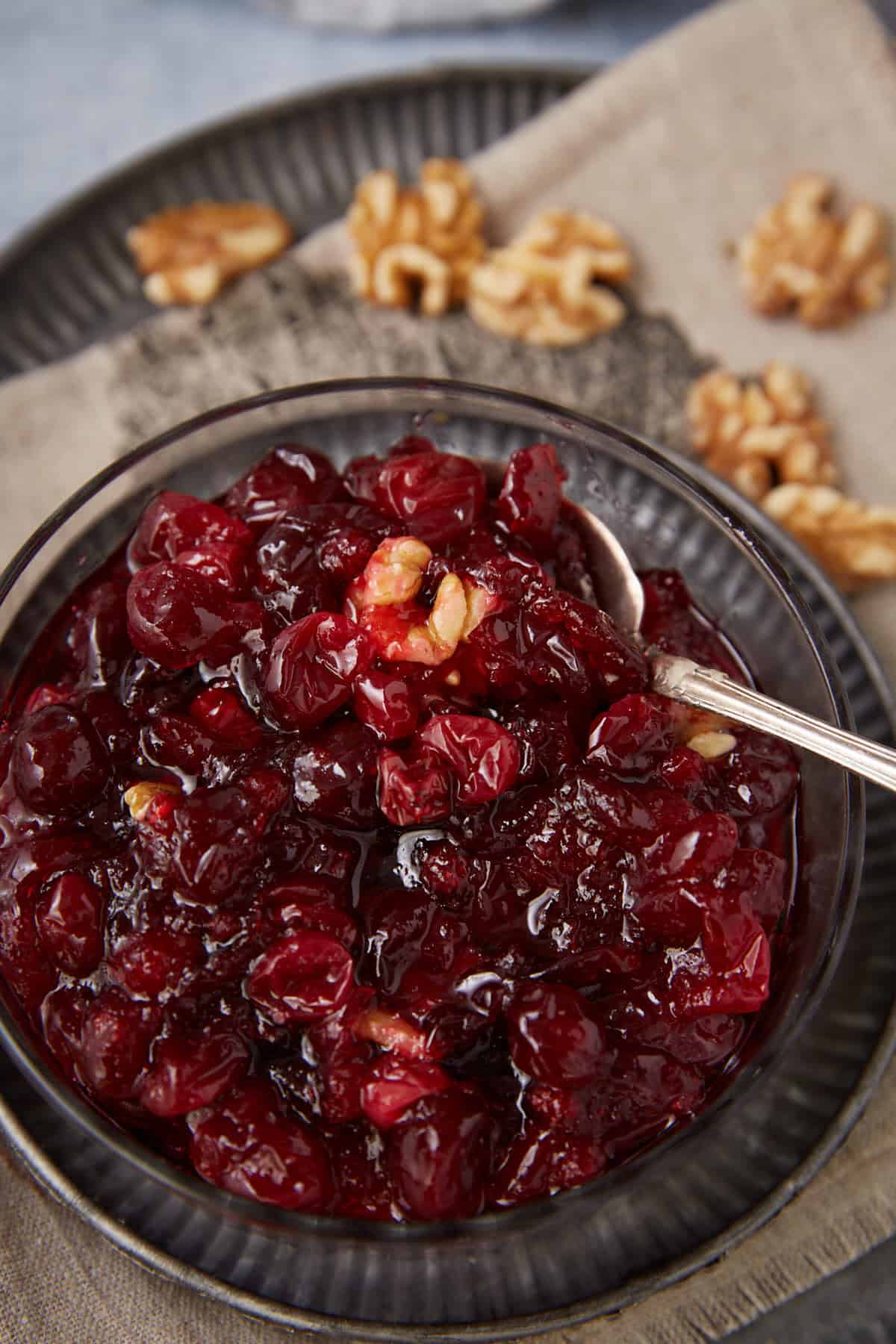 A bowl of chunky homemade cranberry sauce with visible walnut pieces, served with a silver spoon. Walnuts are scattered on a cloth napkin in the background.