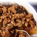 Close-up of Easy Sweet Potato Casserole topped with a buttery, pecan-studded crumble. The golden-brown topping appears crispy and textured. A silver spoon is scooping a portion from the dish, revealing the creamy sweet potato filling beneath.