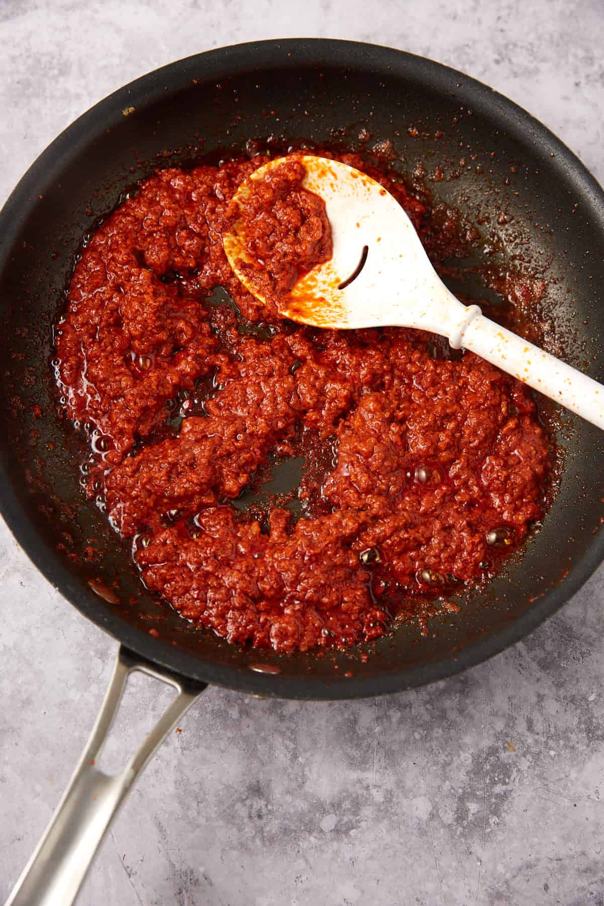 A black frying pan containing a thick, red tomato-based Chorizo Queso Dip being stirred with a white spoon, placed on a light grey textured surface.