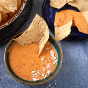 A bowl of creamy Chorizo Queso Dip with a tortilla chip dipped in it, topped with a sprinkle of white powder, sits next to plates of tortilla chips on a blue surface.