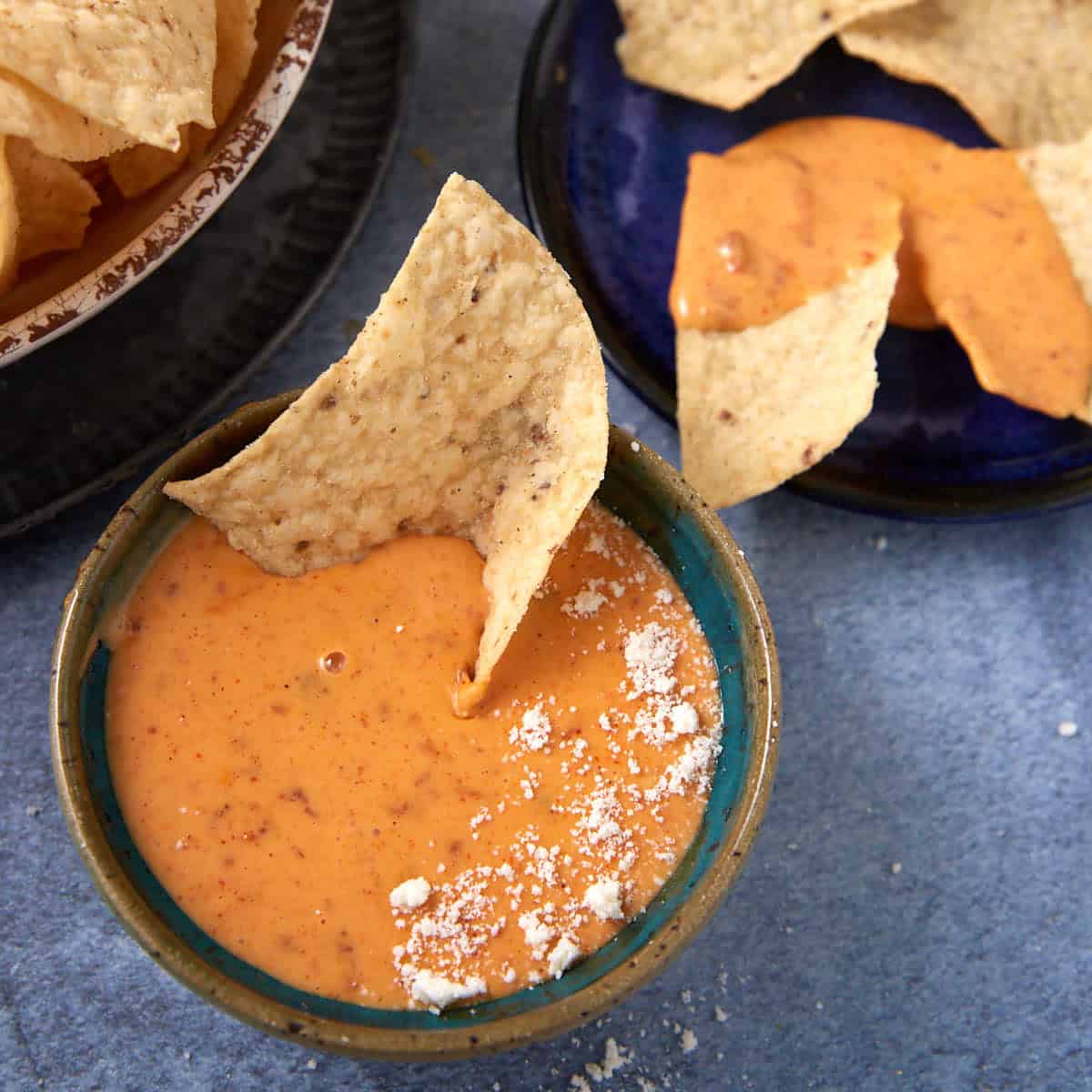 A bowl of creamy Chorizo Queso Dip with a tortilla chip dipped in it, topped with a sprinkle of white powder, sits next to plates of tortilla chips on a blue surface.