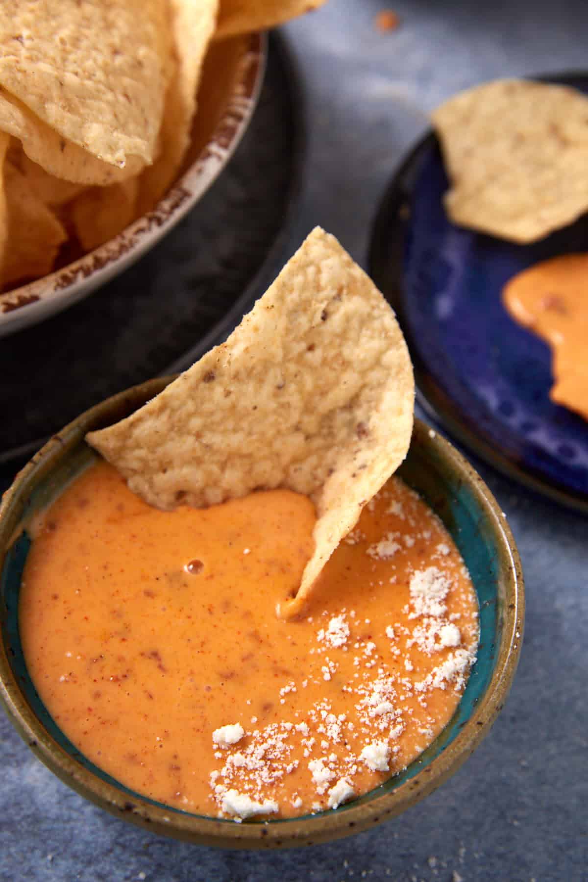 A tortilla chip is dipped into a bowl of creamy orange Chorizo Queso Dip, sprinkled with white cheese. Nearby, more chips are on a plate and in a bowl, perfect for sharing.