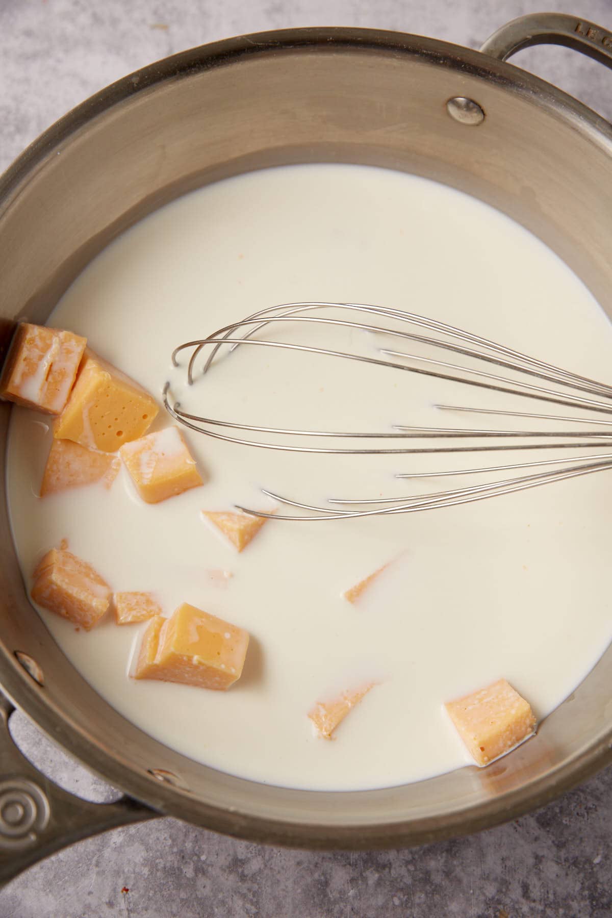 A metal saucepan filled with milk and chunks of cheese for Chorizo Queso Dip is being mixed with a metal whisk on a gray countertop.