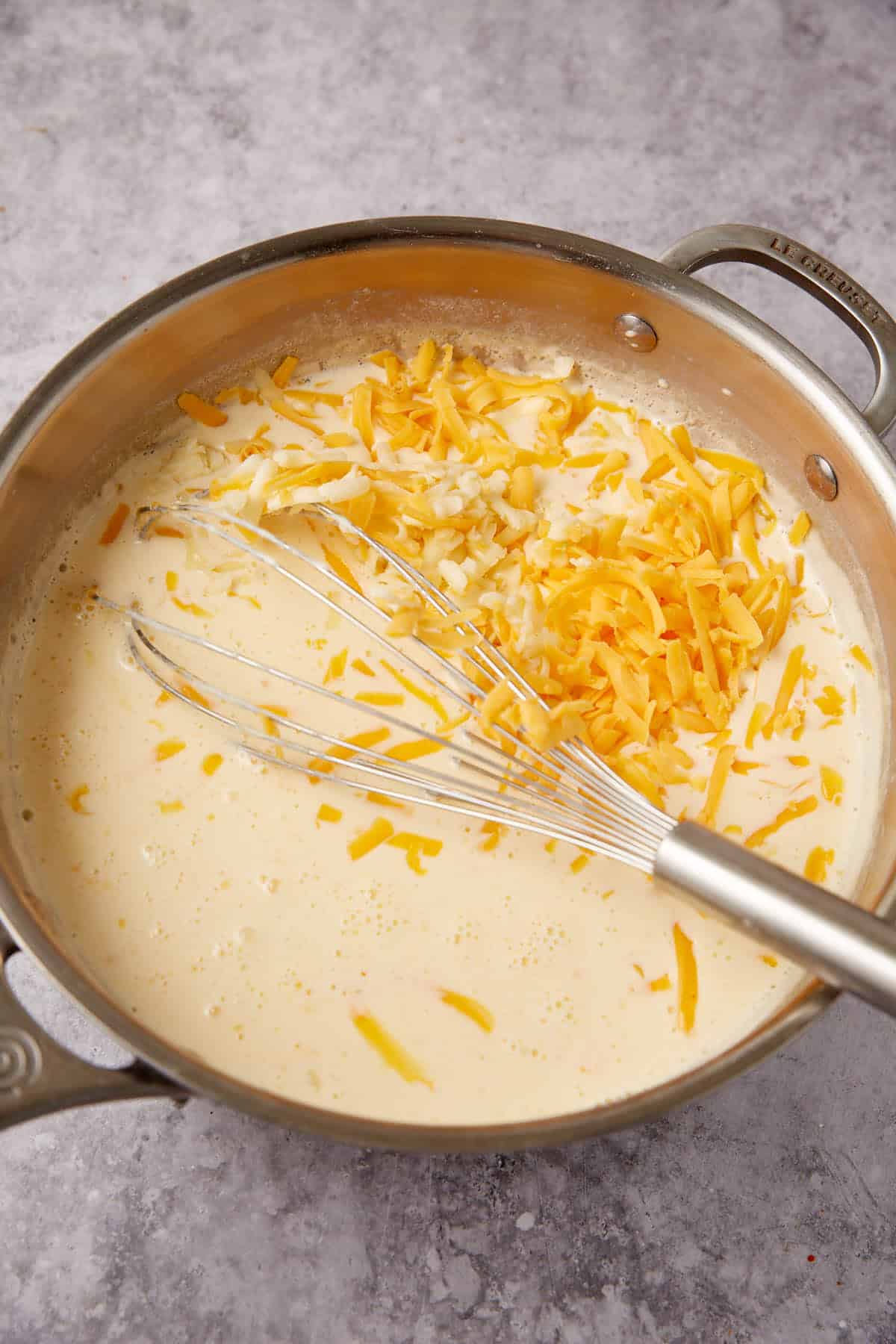 A saucepan filled with creamy milk mixture and shredded cheese for Chorizo Queso Dip is being whisked together on a gray countertop.