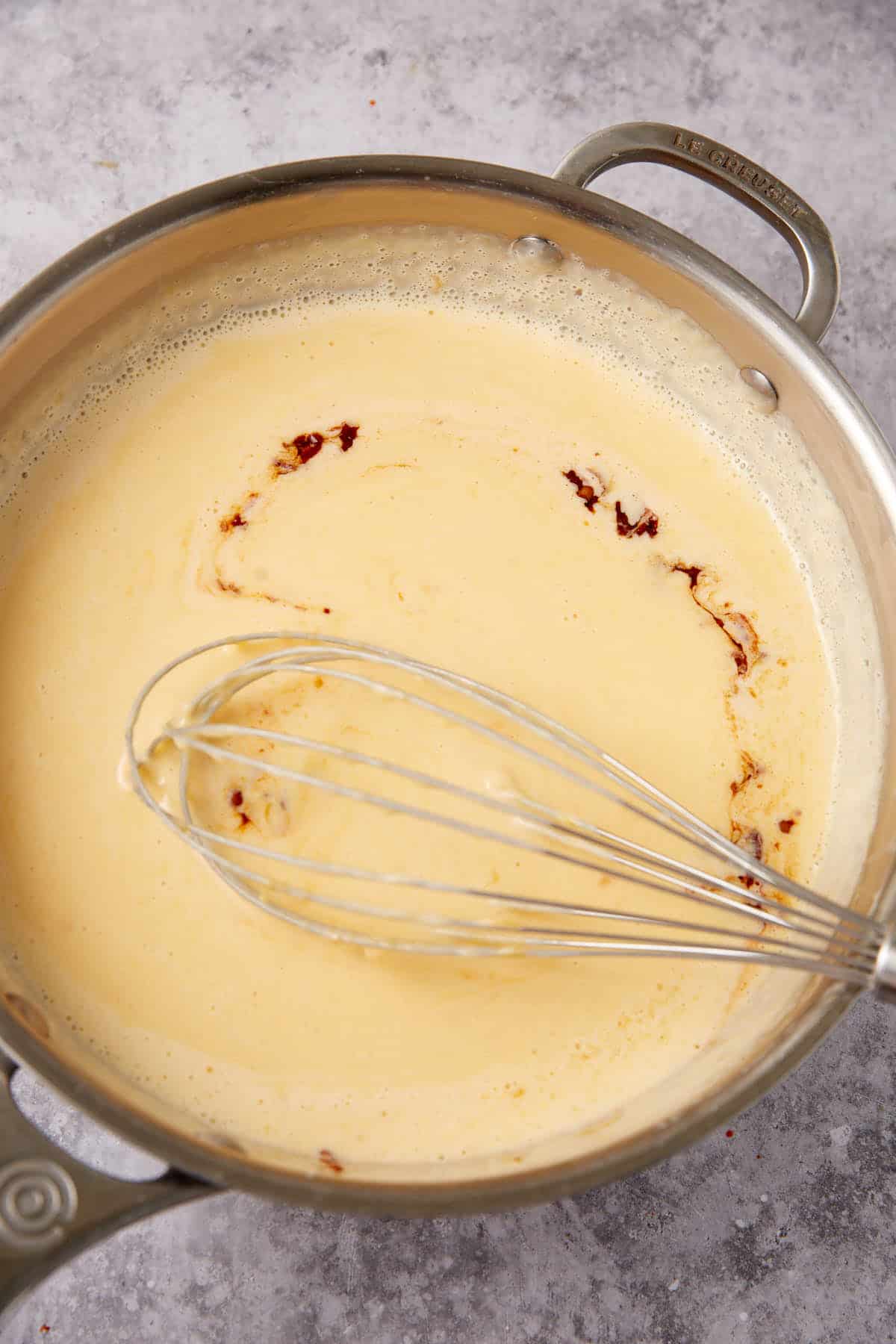 A metal whisk stirring a creamy yellow Chorizo Queso Dip with some brown seasoning in a stainless steel saucepan on a gray countertop.