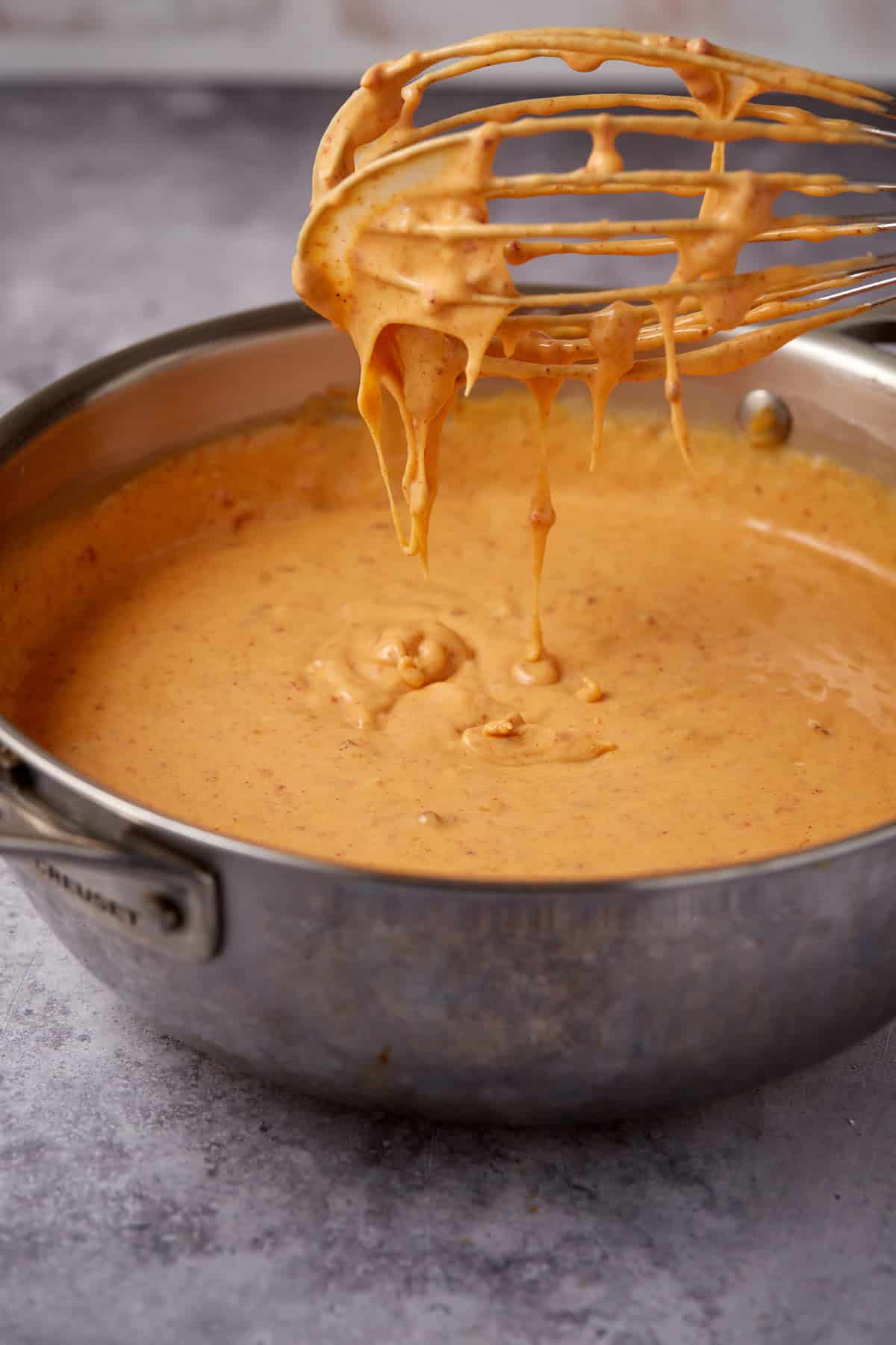 A metal whisk covered in creamy orange Chorizo Queso Dip is held above a saucepan filled with more of the savory cheese sauce on a gray countertop.