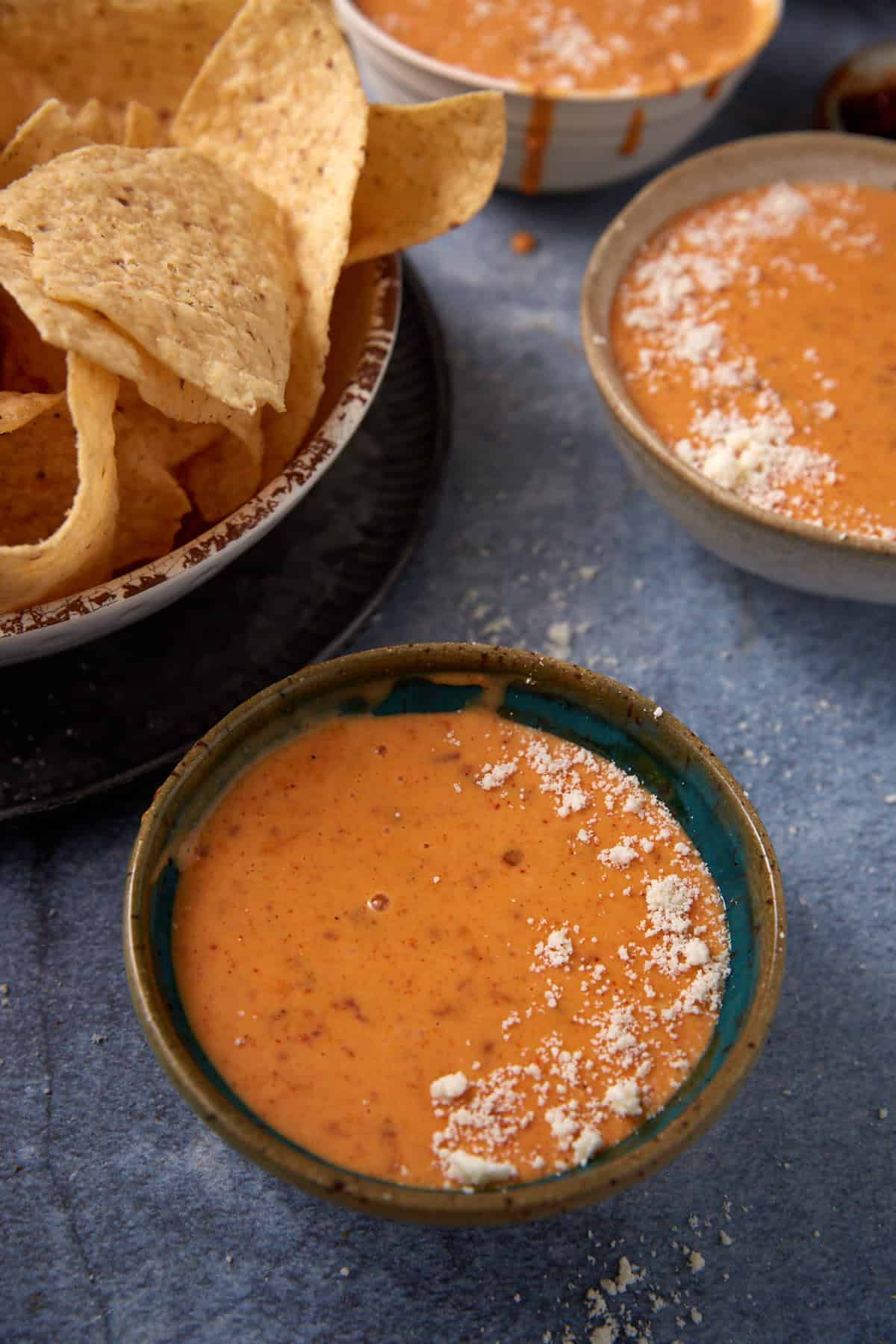 Bowls of creamy orange Chorizo Queso Dip, some topped with crumbled cheese, sit on a blue surface next to a bowl filled with tortilla chips.