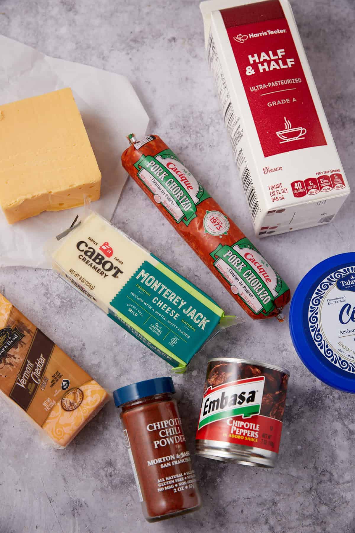 Assorted ingredients on a gray surface for Chorizo Queso Dip, including cheddar, Monterey Jack cheese, half & half, two pork chorizo sausages, chipotle chili powder, canned chipotle peppers, a cheese wedge, and sour cream.