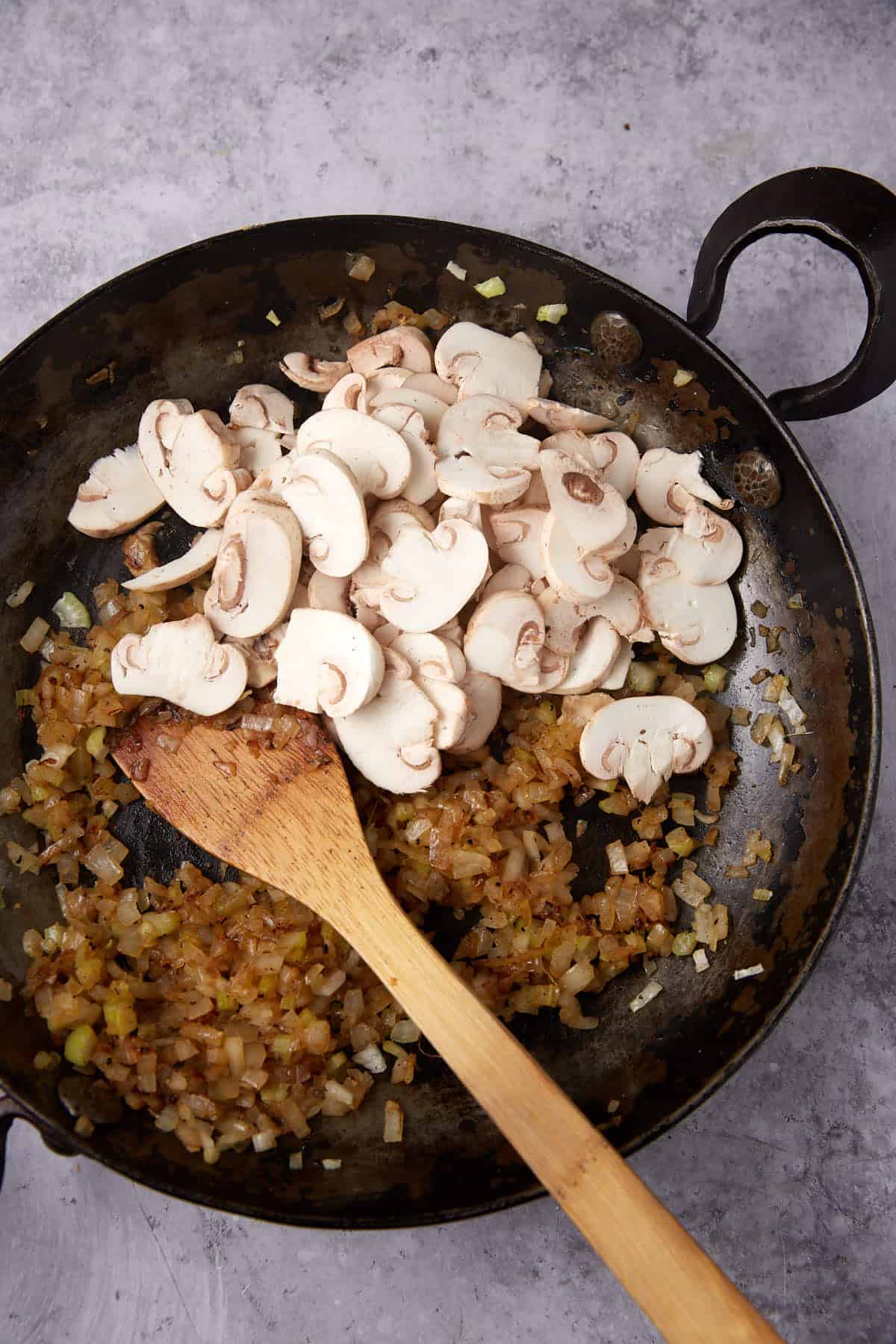 mushrooms being added to the sautéed celery and onions in a skillet with a wooden spoon.
