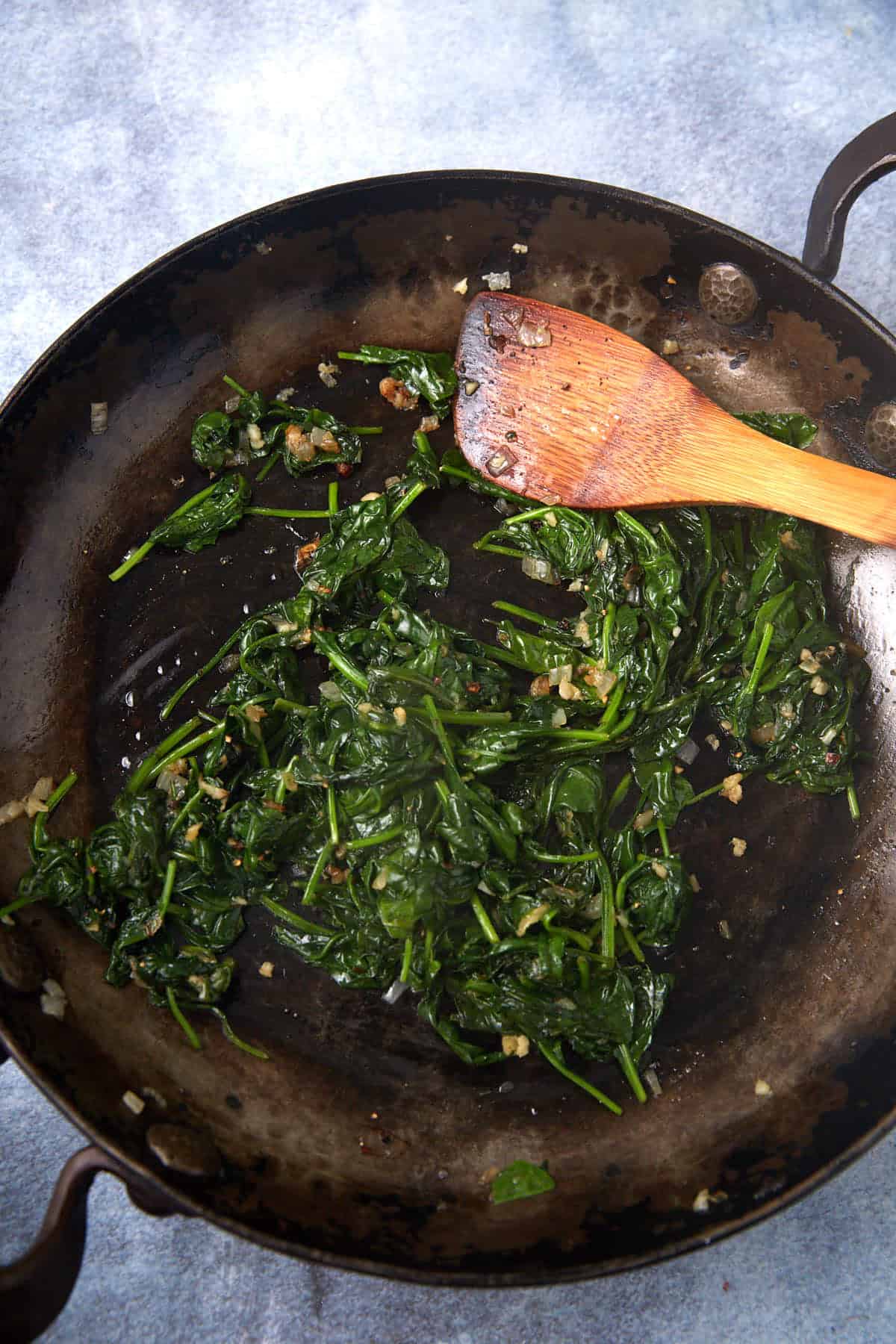 Sautéed spinach with garlic and onions in a dark skillet, perfect as a base for an easy spinach artichoke dip, with a wooden spatula resting on the side of the pan.