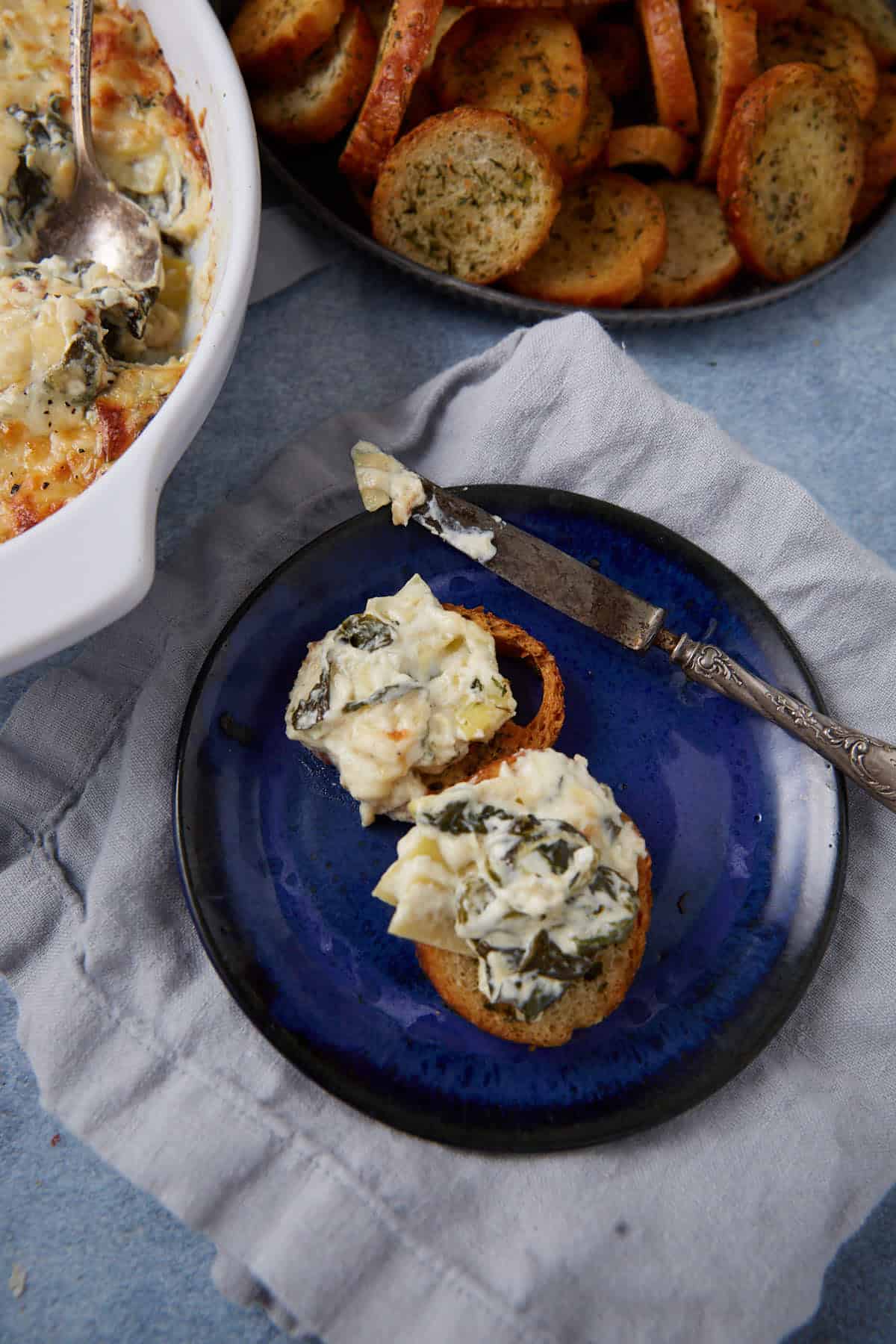 A blue plate holds two toasted bread slices topped with easy spinach artichoke dip. A knife with some dip rests next to them. In the background, a bowl of dip and a plate of toasted bread slices are visible.
