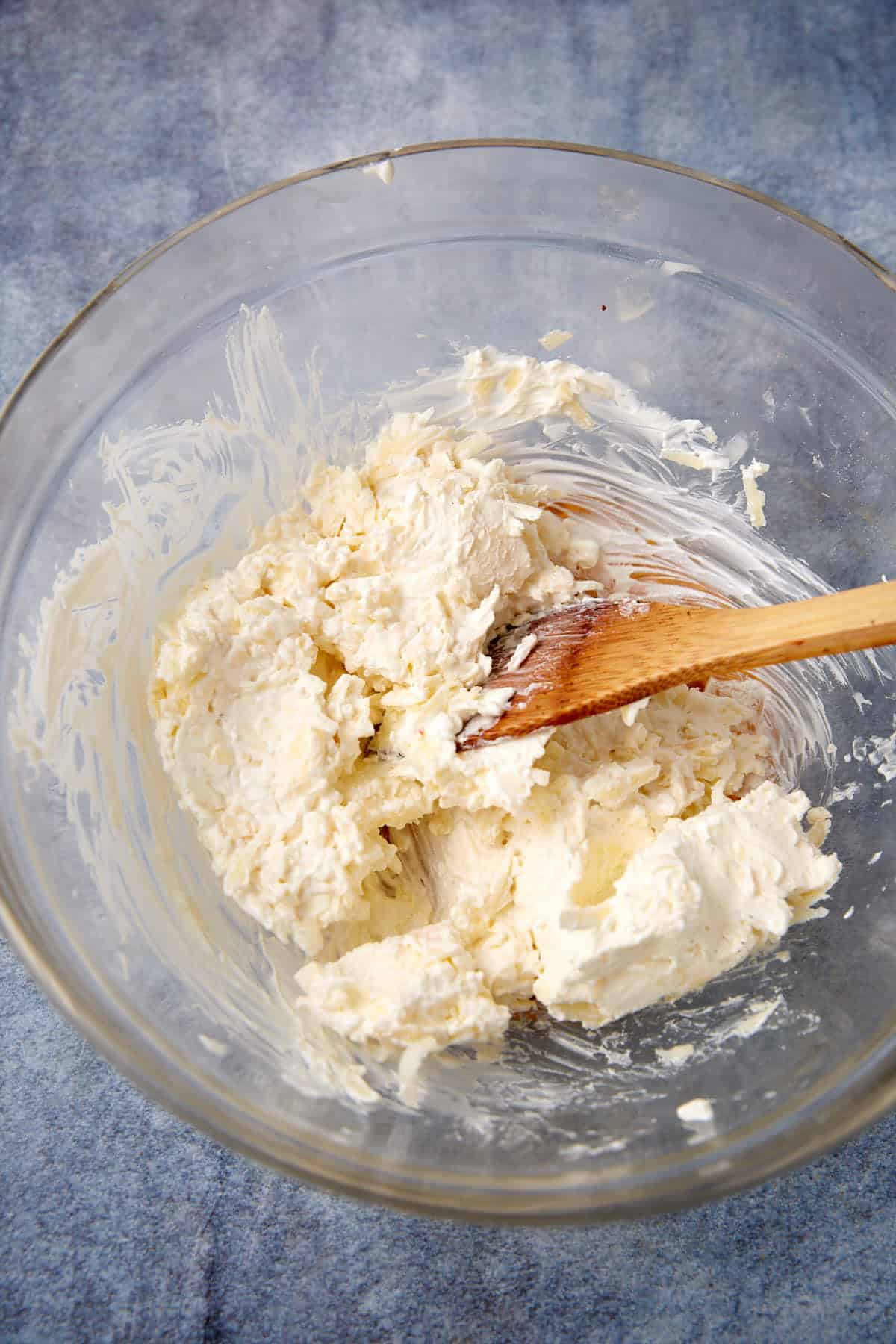 A clear glass bowl with a creamy, white cheese mixture for easy spinach artichoke dip is being stirred by a wooden spoon, set on a textured blue-gray surface.