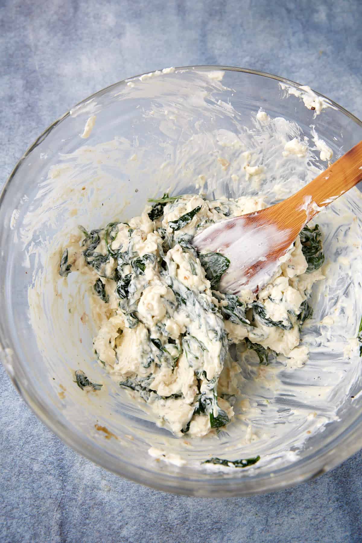 A glass bowl filled with an easy spinach artichoke dip sits on a blue surface, its creamy spinach and cheese mixture inviting. A wooden spoon coated in the dip rests inside the bowl.