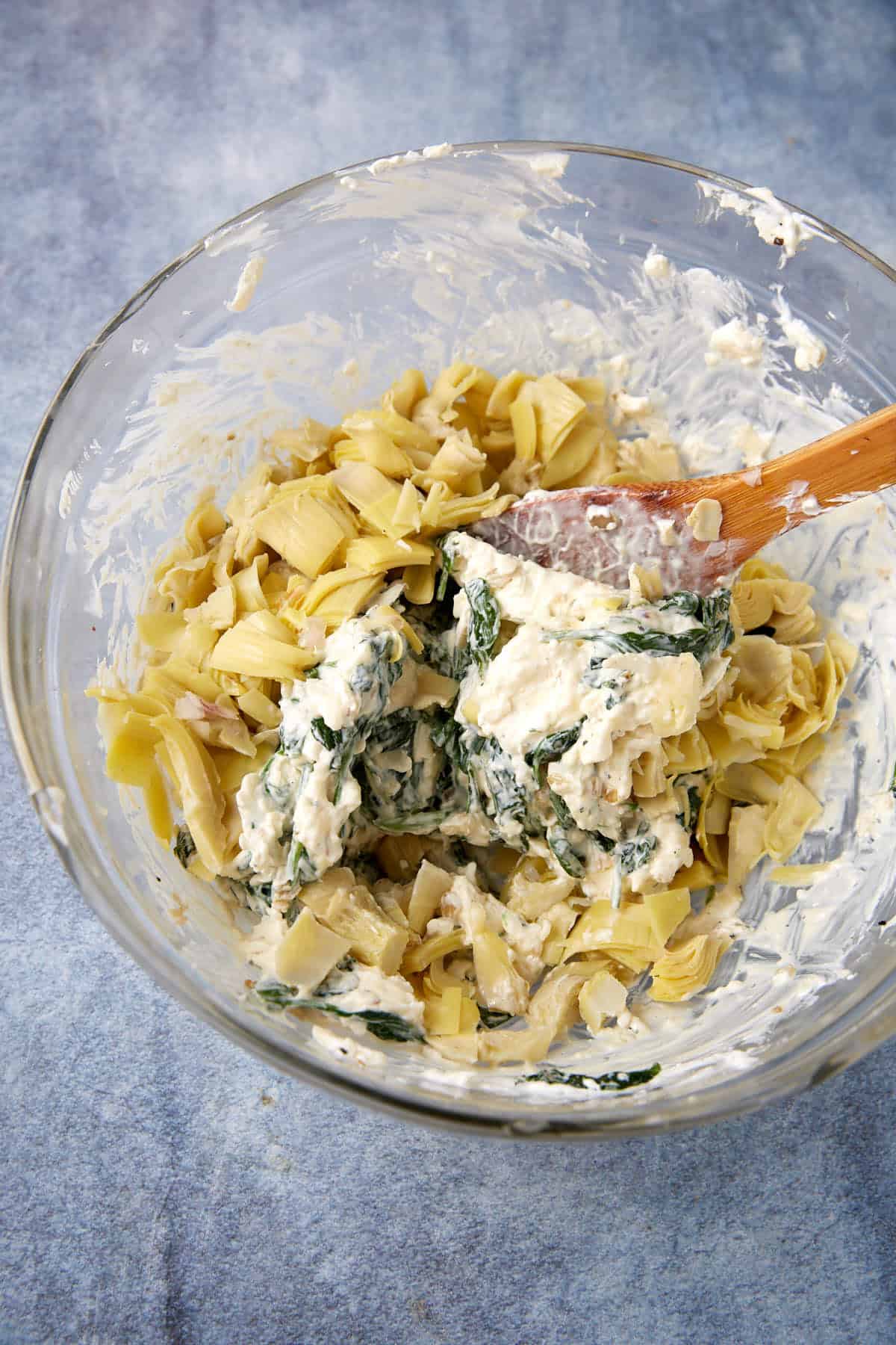 A glass bowl containing easy spinach artichoke dip—chopped artichokes and creamy spinach mixture being stirred with a wooden spatula—set on a blue surface.